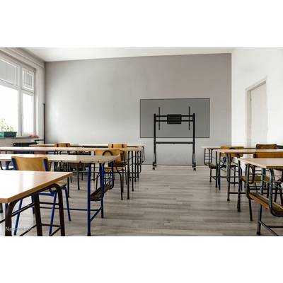 An empty classroom with chairs and tables. A screen holder is mounted on the grey wall in the background. Windows are located on the left side.