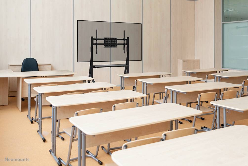 A vacant, bright classroom with wooden tables and chairs. In the background, a large display stands on a stand in front of a wall.
