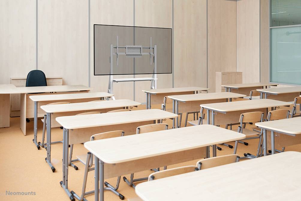 Classroom with empty desks and chairs. At the front, a teacher's desk with a chair, and a display stand in the background.