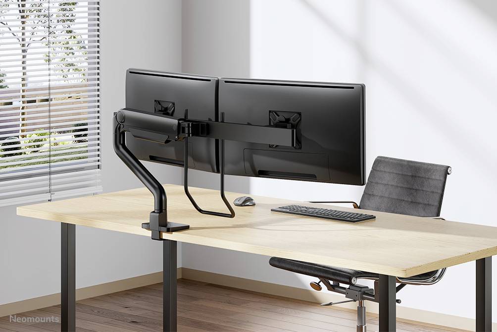 A desk with two monitors mounted on a monitor arm, alongside a keyboard and a mouse. A chair stands beside it.