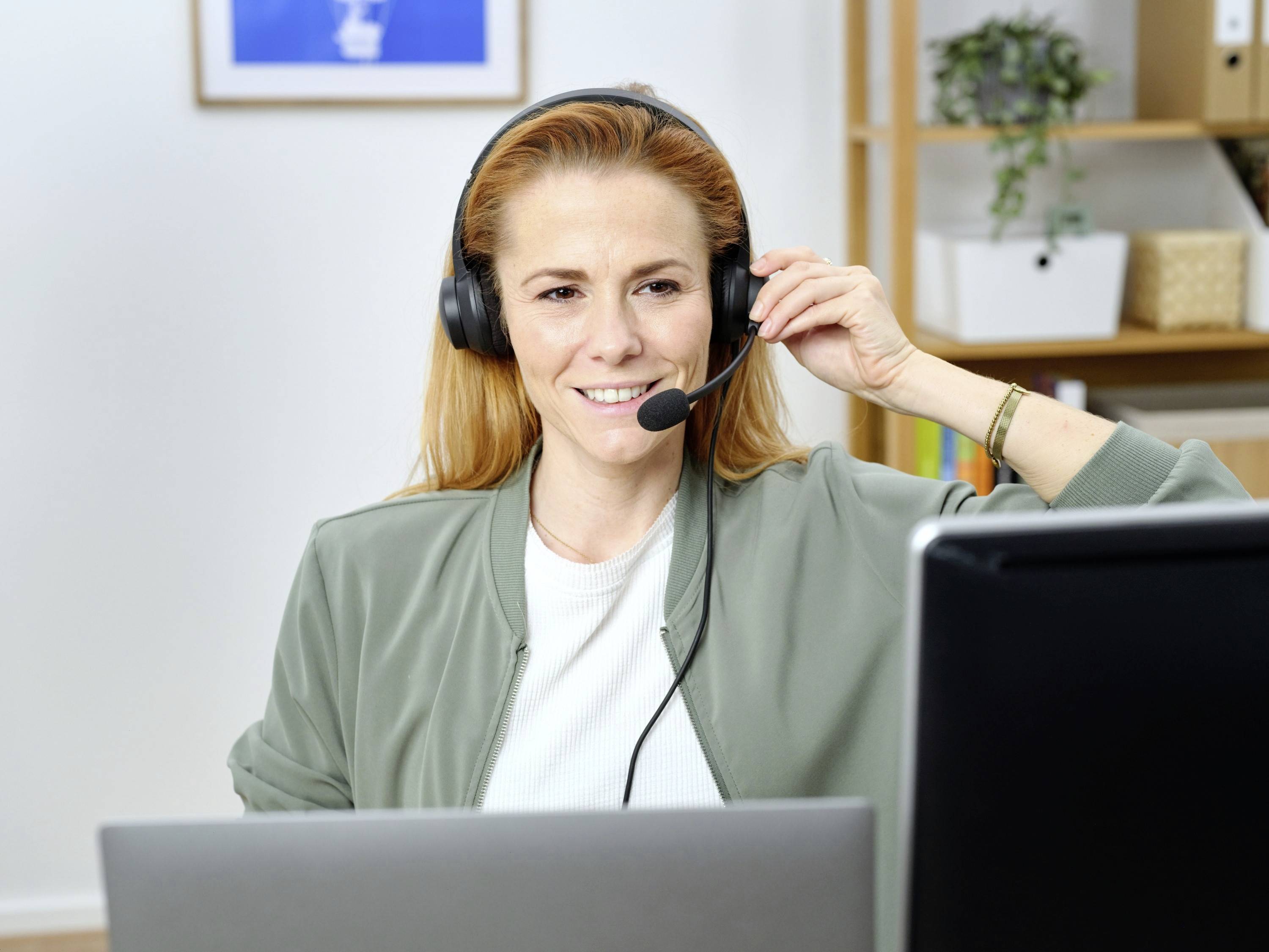 A woman wearing headphones and a microphone sits in front of a computer screen, smiling, indicating a customer service or remote working context.