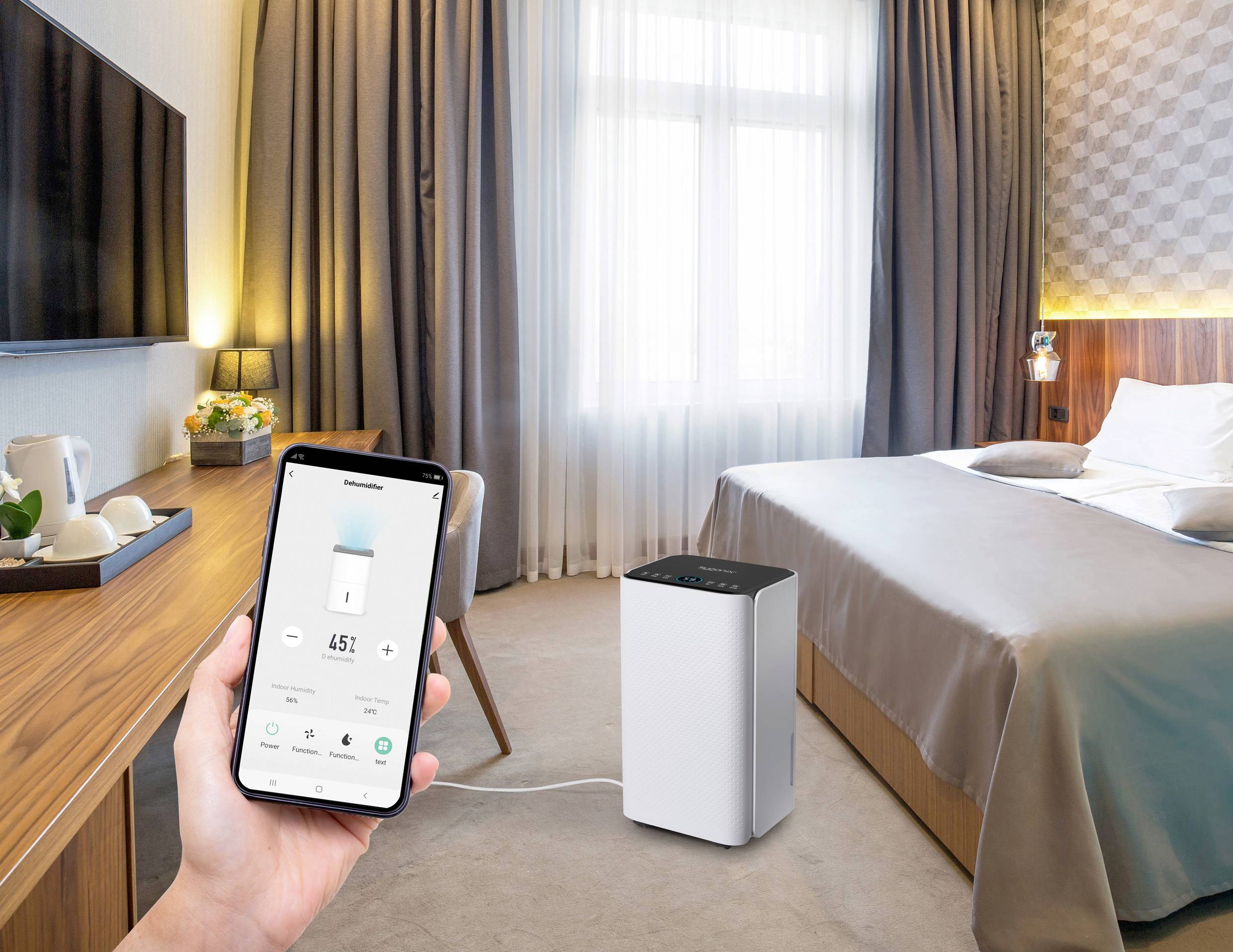 A hotel room with a bed and an air purifier on the floor. A hand is holding a smartphone displaying the air purifier's controls.