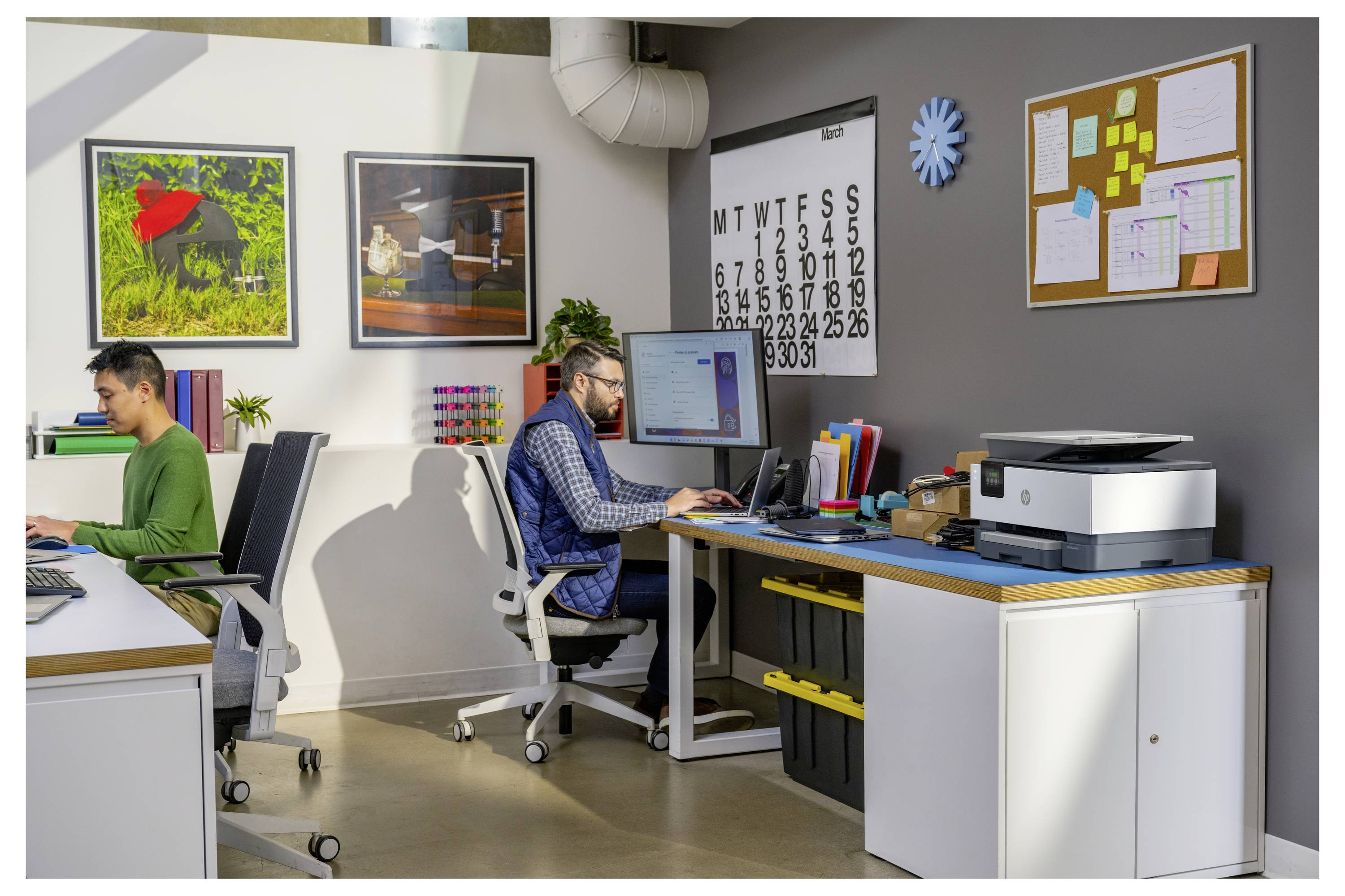 Two men work at desks in a shared office, each using a computer beside printers, wall art, a calendar, and a bulletin board.