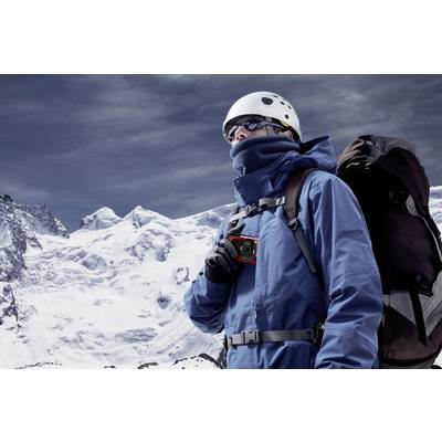 A mountaineer in warm gear stands in front of snow-covered mountains, holding a compass and carrying a large rucksack.