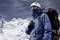 A mountaineer in warm gear stands in front of snow-covered mountains, holding a compass and carrying a large rucksack.