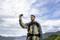 A man in outdoor clothing takes a selfie with his mobile phone against a backdrop of mountains and cloudy sky.