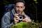 A man with a rucksack is photographing a small plant on a moss-covered stone in the woods.