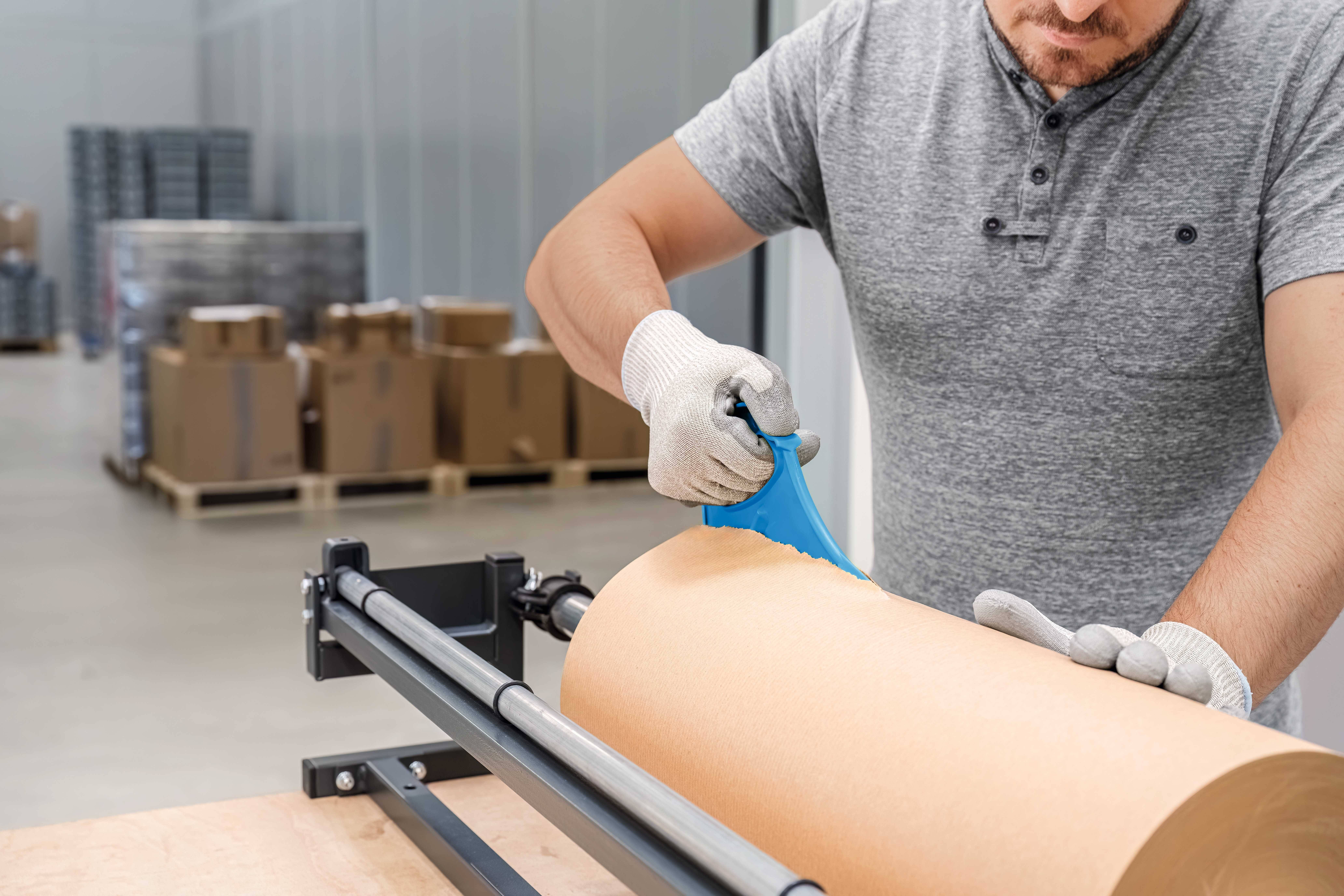 A man in a grey shirt and gloves is cutting paper on a machine in a warehouse with boxes in the background.