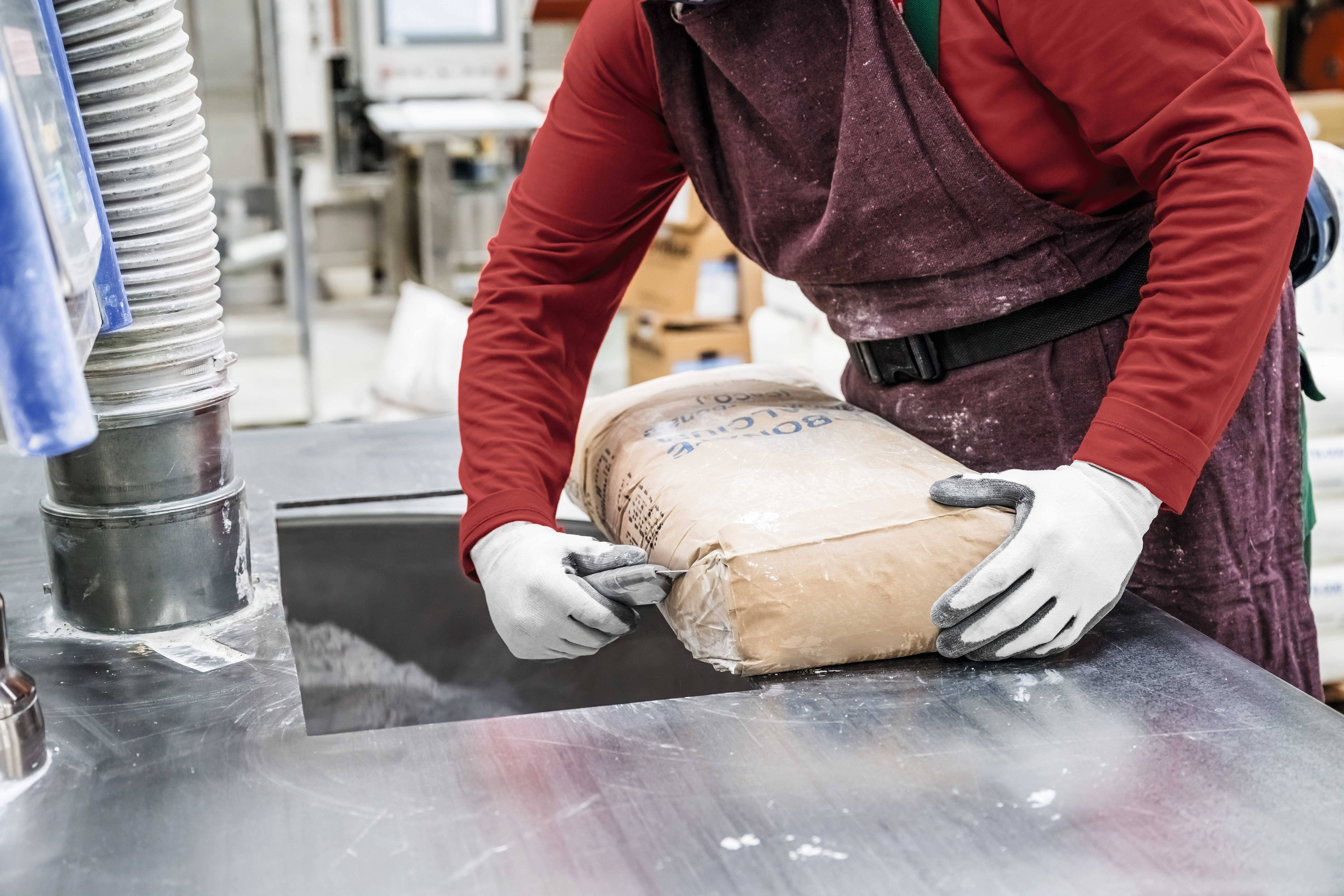 A person wearing work attire tips a sack of flour into a machine in a food processing facility.