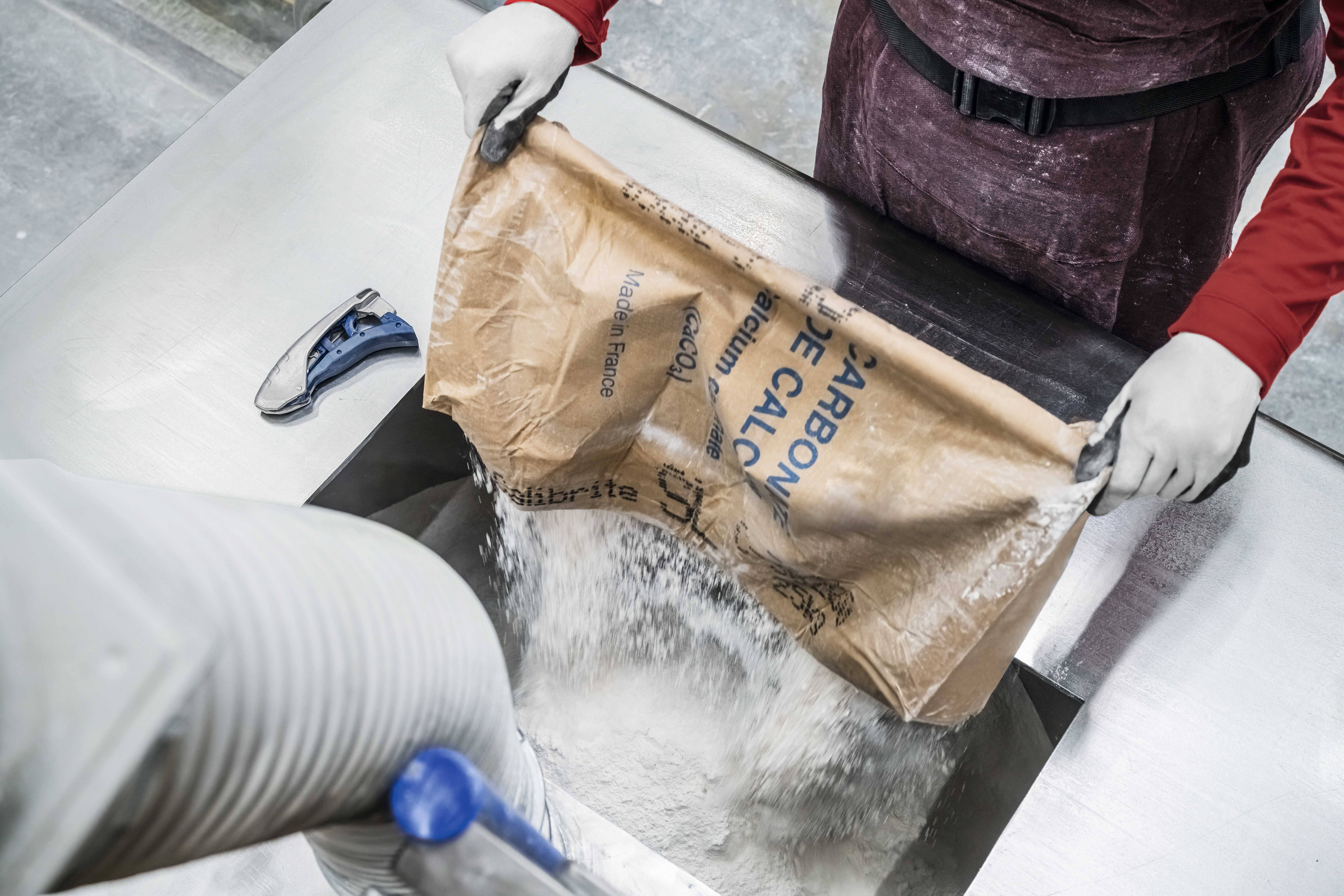 A person wearing gloves is emptying a paper sack of lime powder into an industrial machine. A handheld device lies beside it.