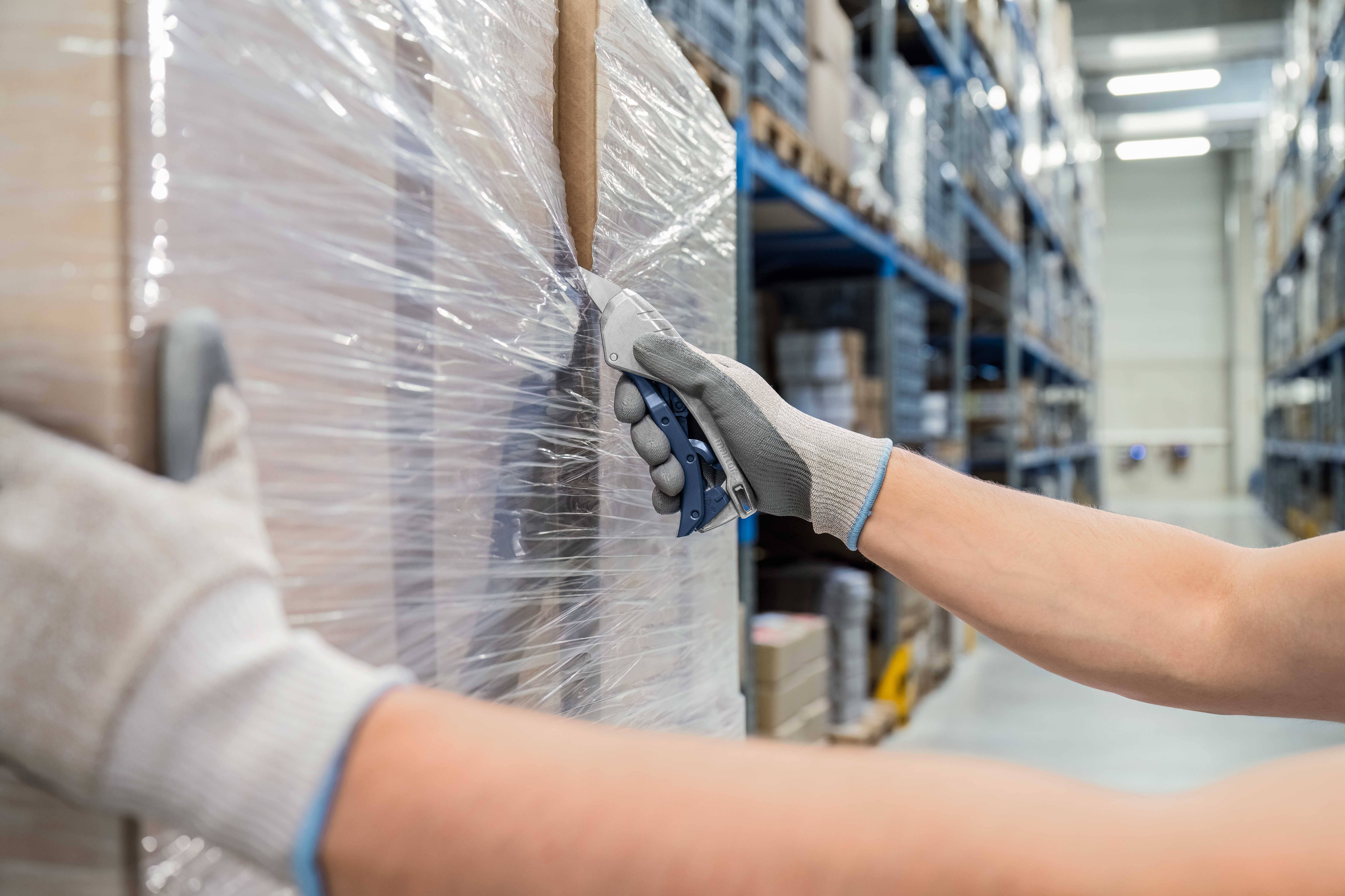 A person cuts plastic film from a pallet in a warehouse while wearing work gloves. Shelves full of boxes are in the background.