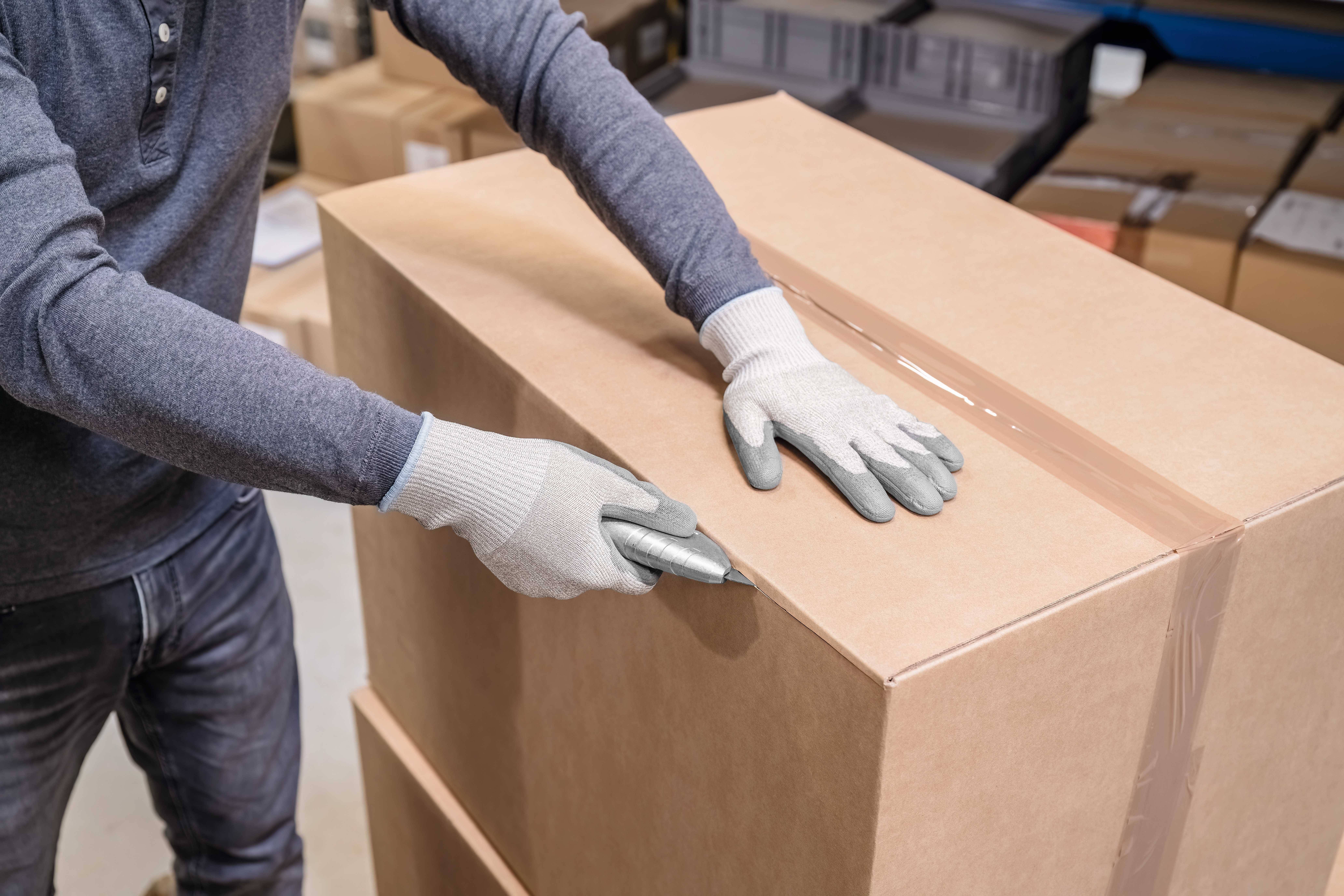 A person wearing blue clothing opens a large cardboard box with a knife. Paper boxes in the background indicate a warehouse setting.