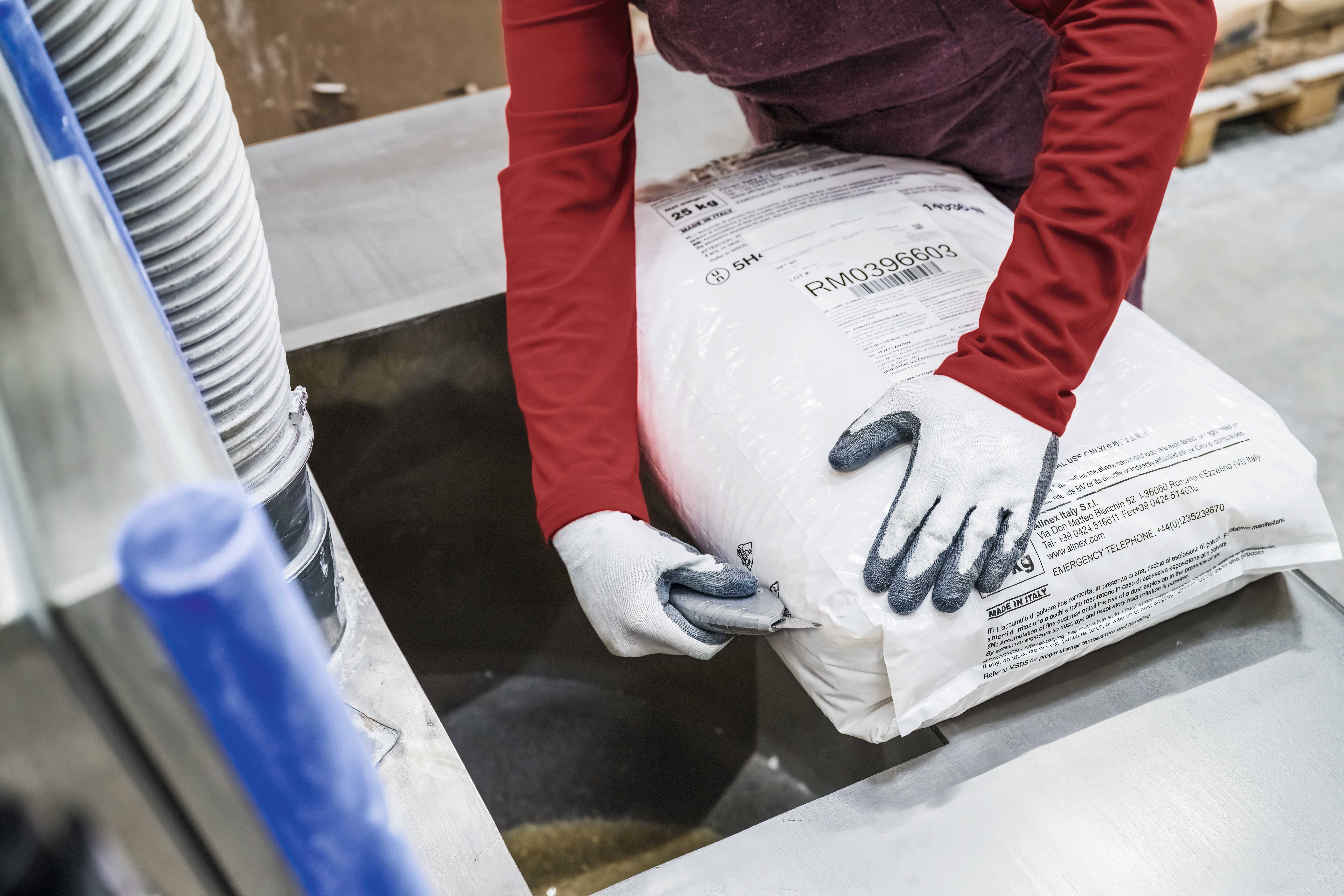 A person wearing gloves is emptying a sack of material into a funnel in an industrial setting.