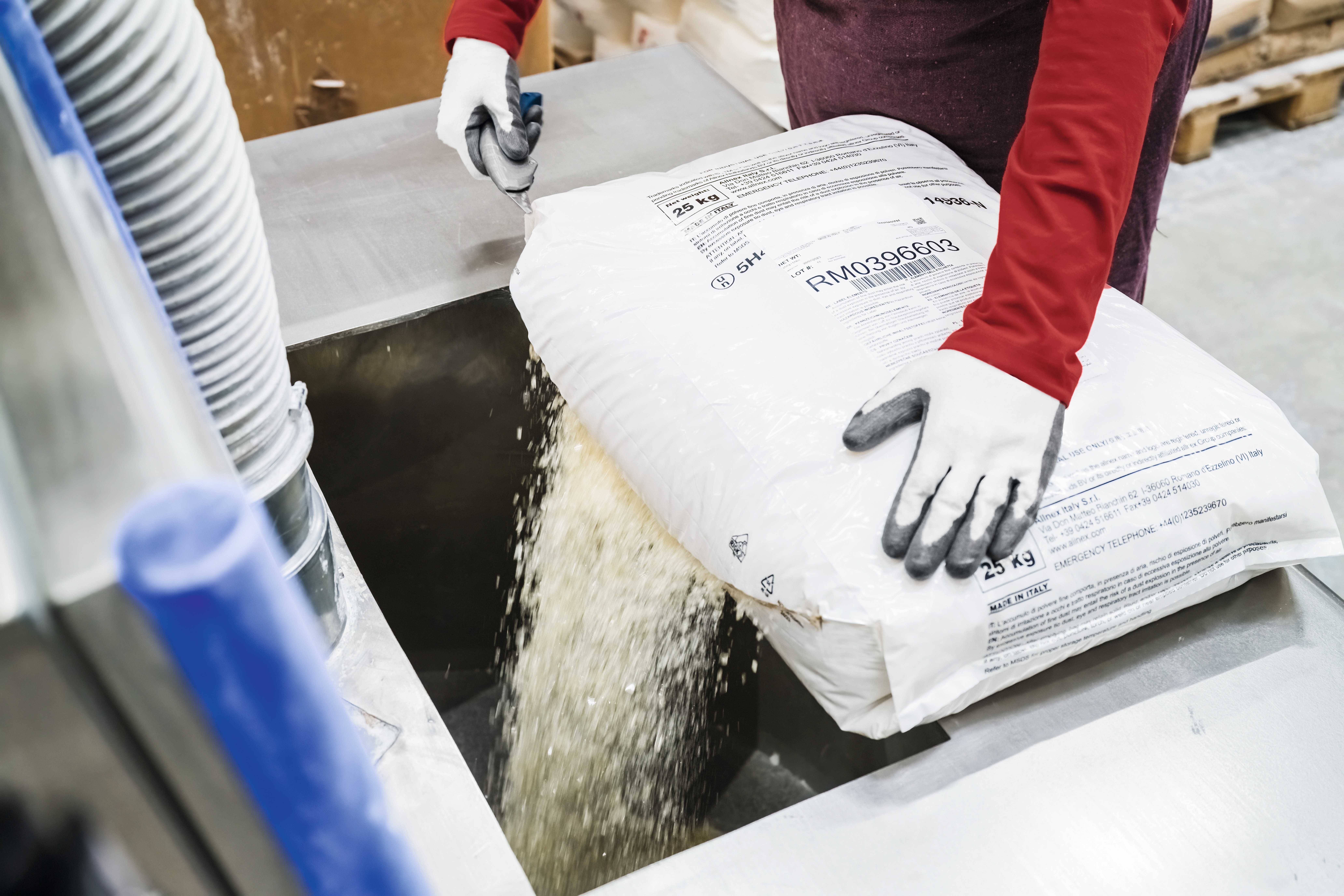 A person is pouring granules from a large, white bag into a machine. The person is wearing red clothing and white gloves.