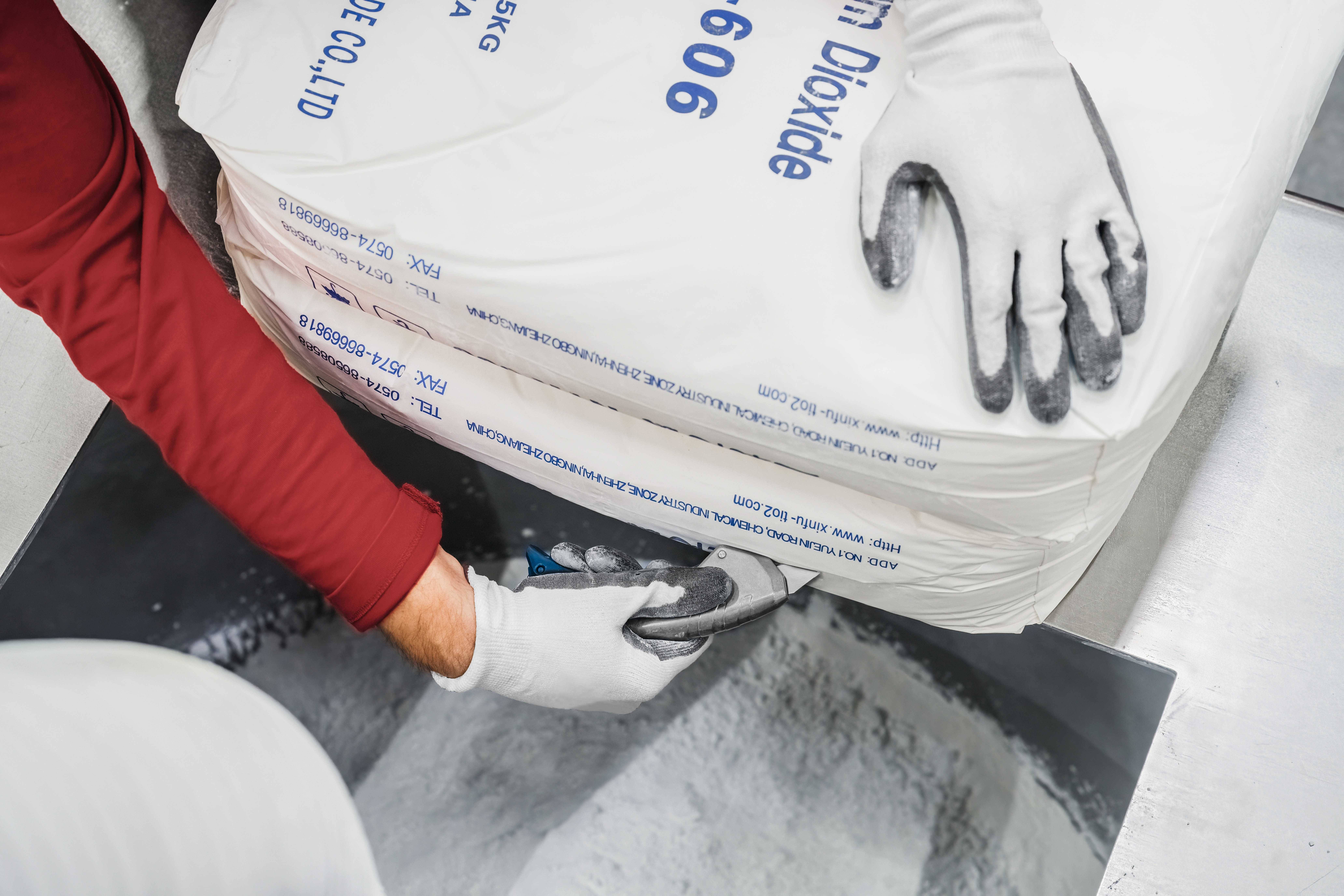 A person in red sleeves opens a large sack of white powder on a scale. The hand is wearing white gloves.