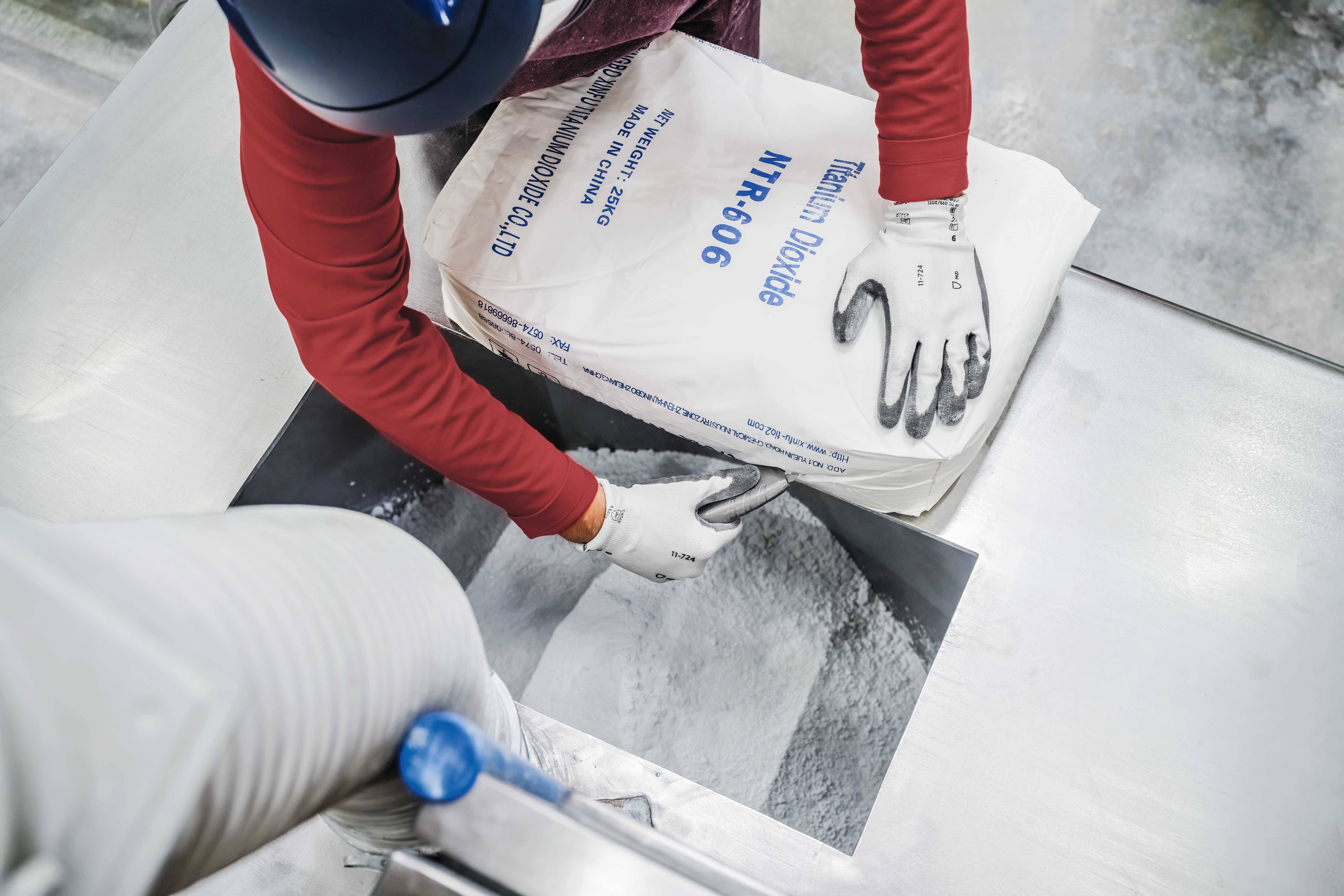 A person wearing gloves is pouring titanium dioxide from a labelled sack into a machine. Industrial environment.