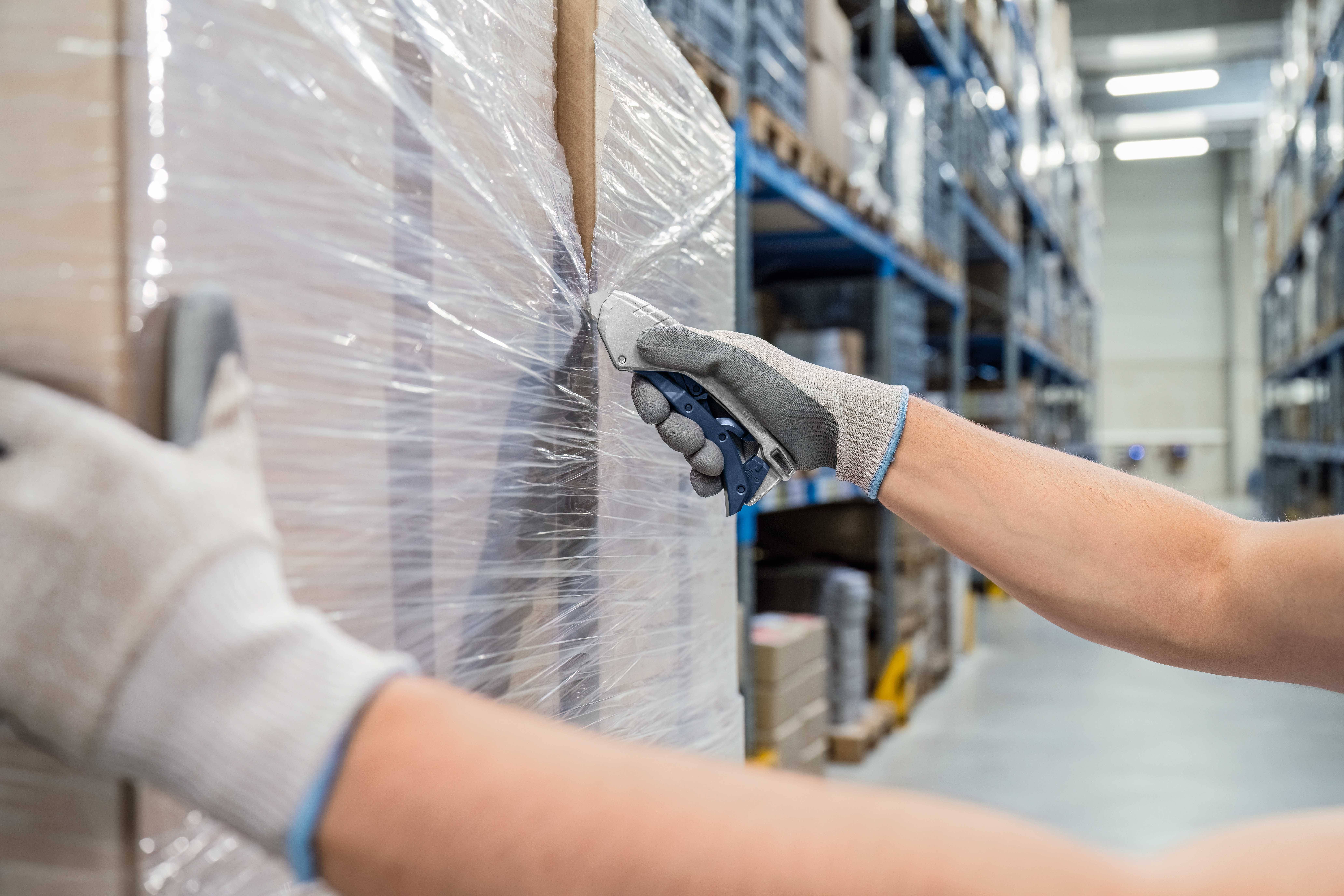 A person wearing gloves is cutting film from a pallet in a large warehouse. Shelves with cardboard boxes are visible in the background.