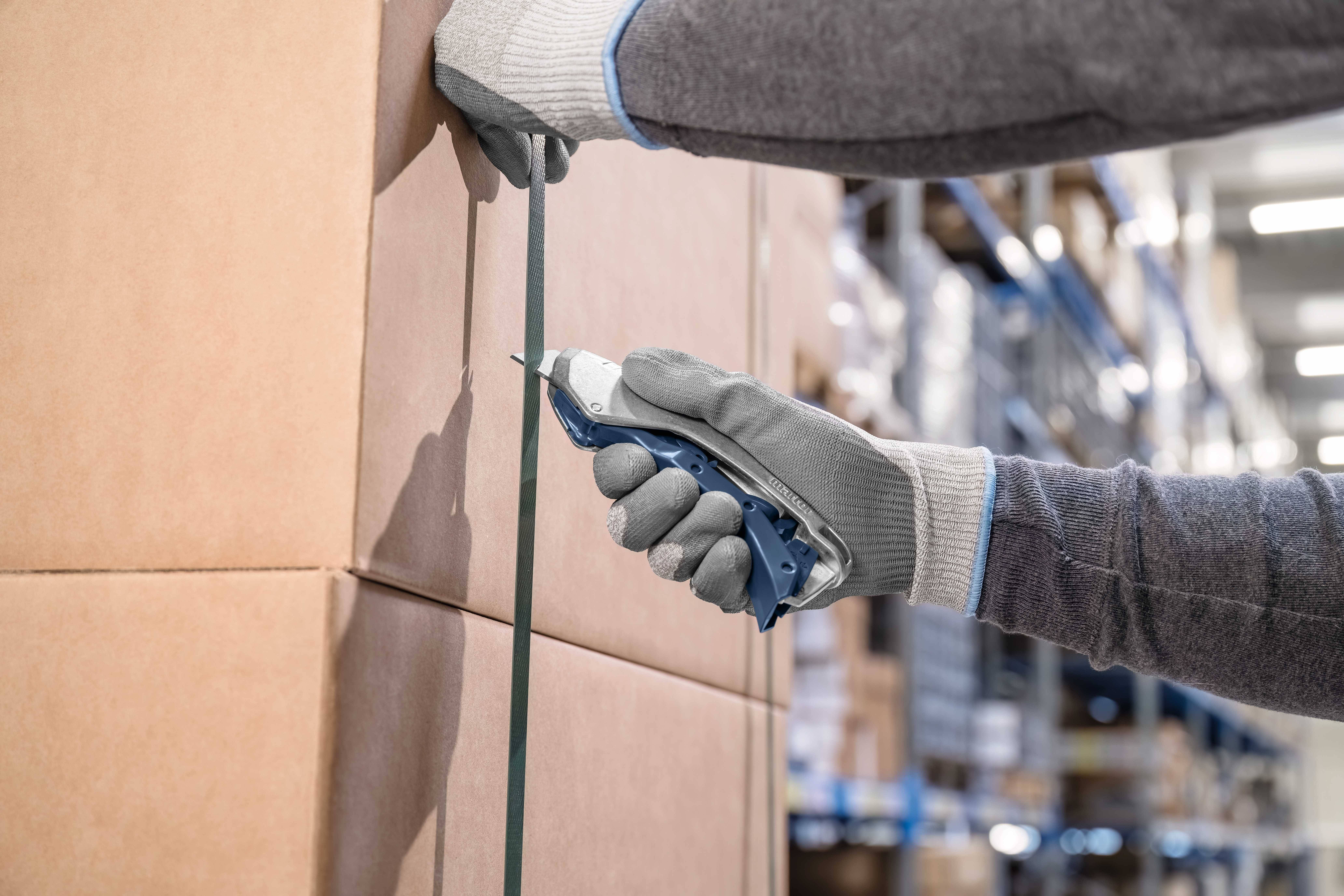 A worker pulls plastic tape over large cardboard boxes in a warehouse, wearing gloves.