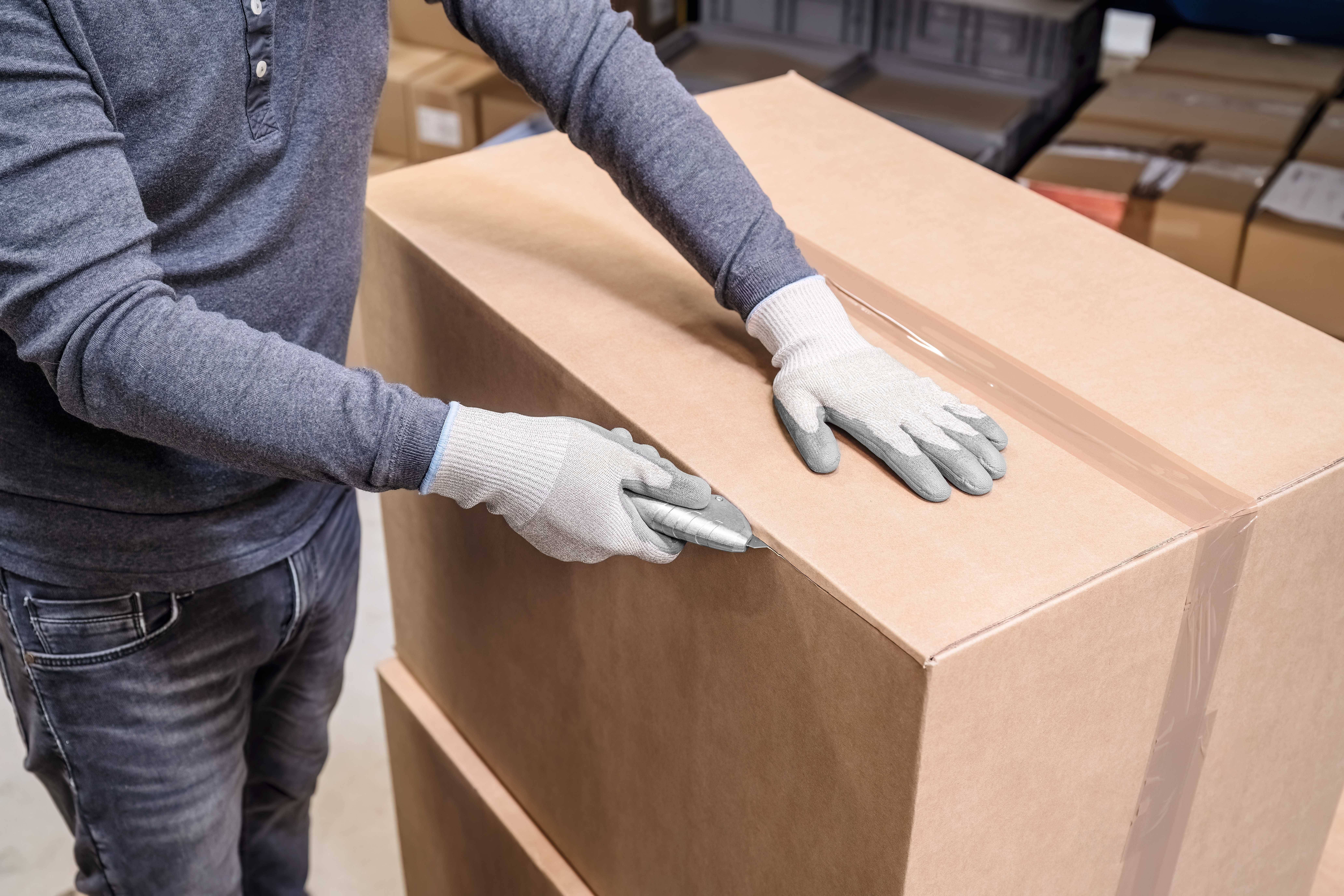 A person wearing grey gloves is opening a large cardboard box with a knife in a storage room.