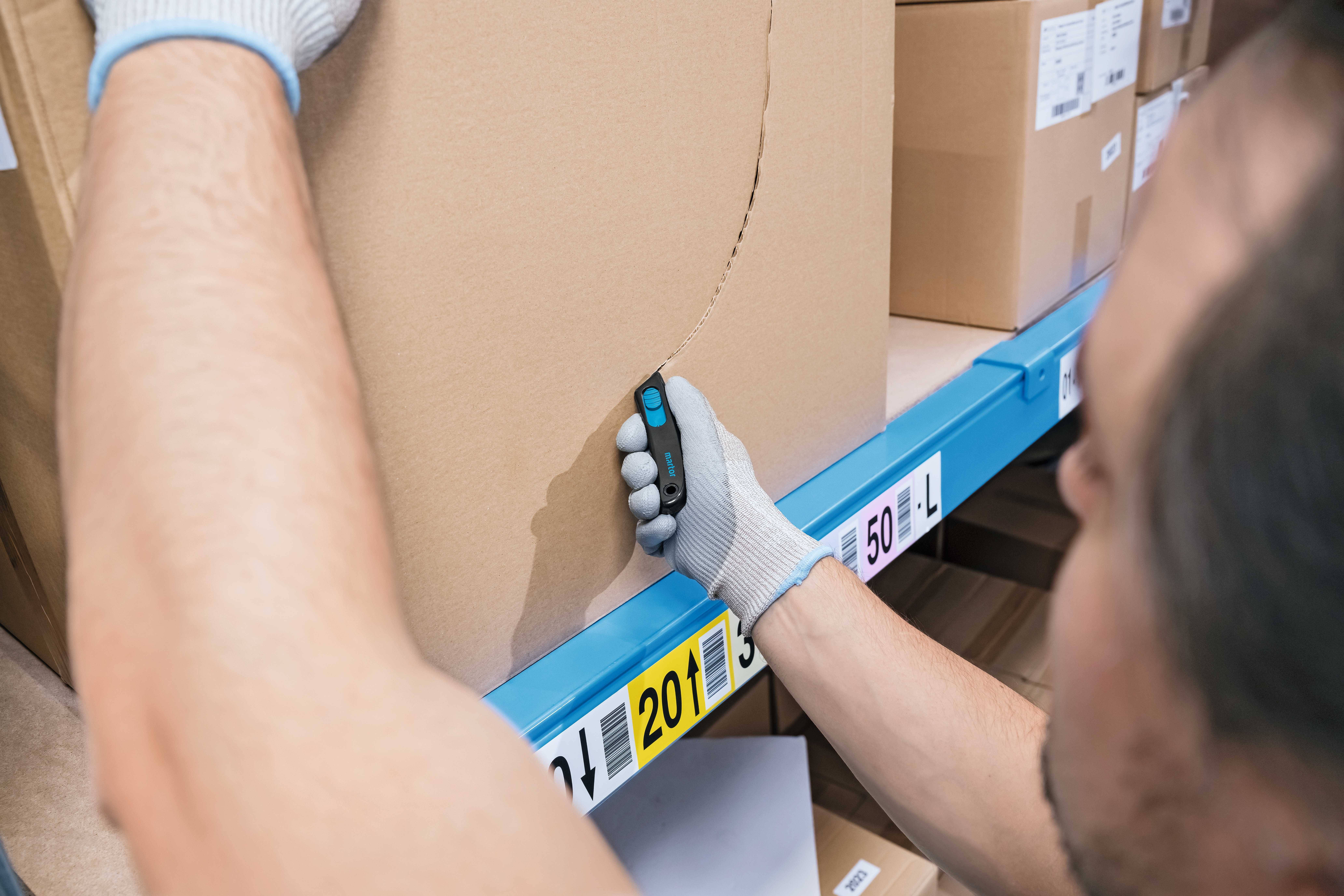A person carefully opens a cardboard box using a craft knife. In the background, additional boxes can be seen on a shelf.