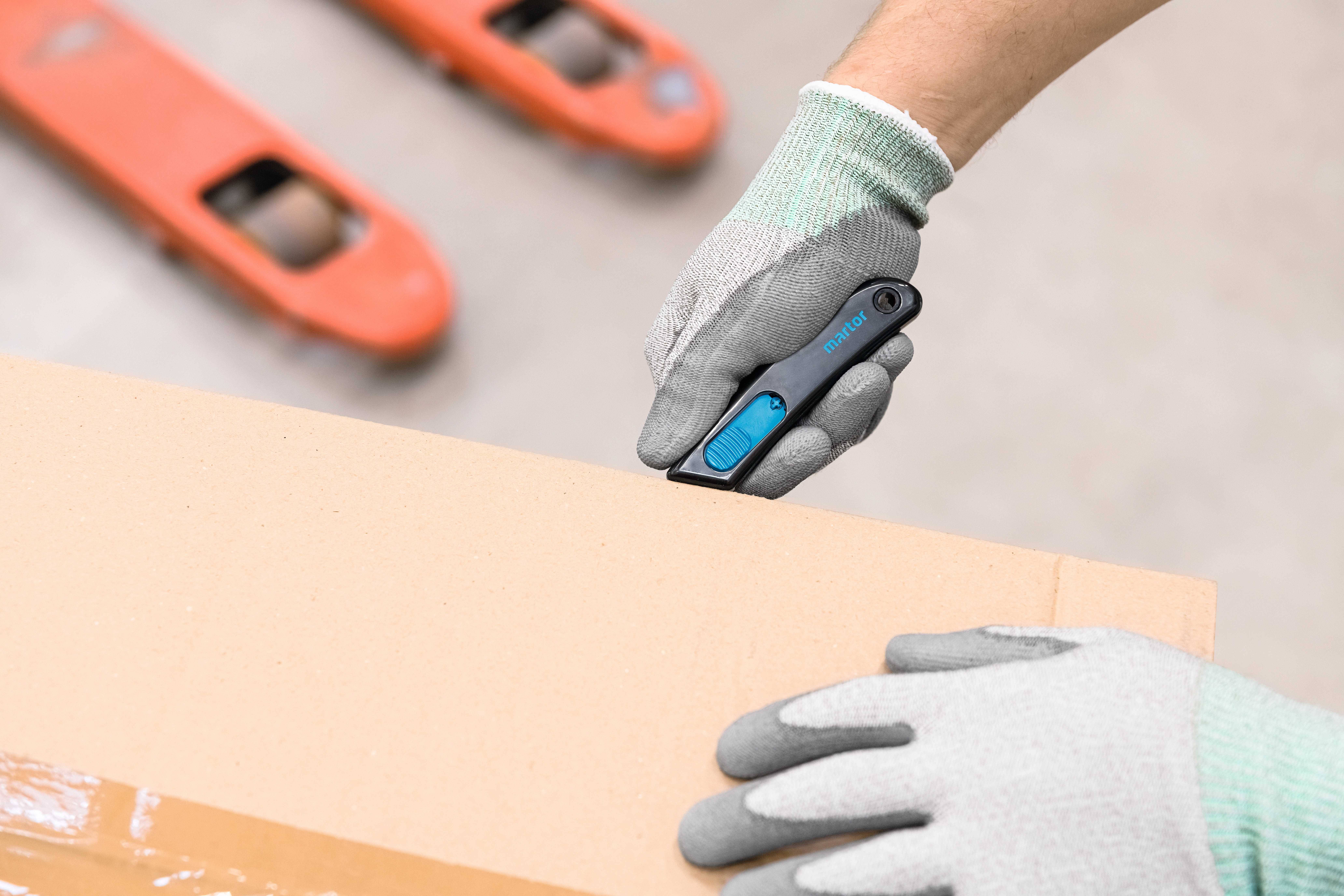 A person is opening a cardboard box using a blue utility knife and grey gloves. Two red pallets are visible in the background.