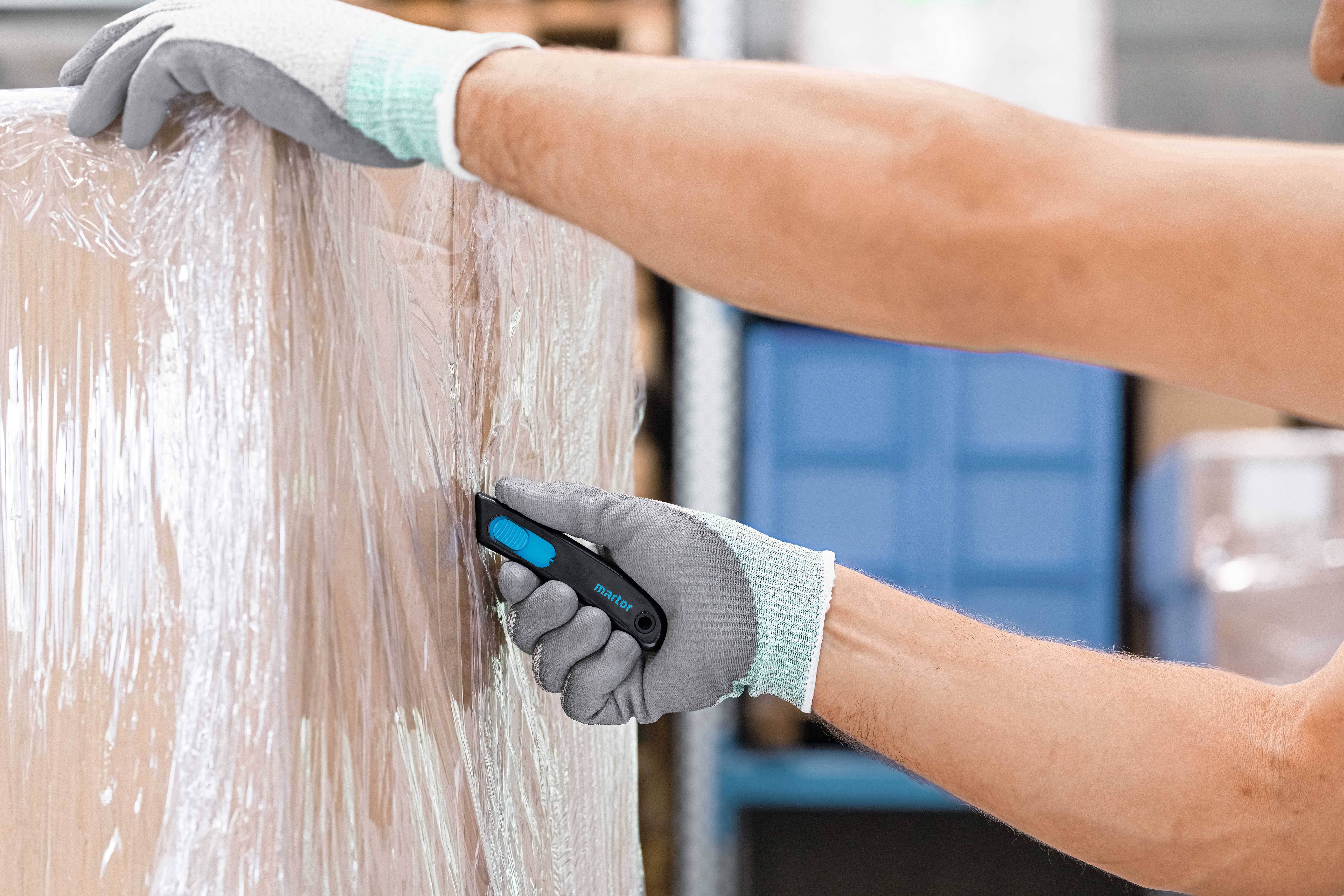 A person wearing gloves is cutting plastic film from a pallet with a knife. Cardboard boxes are stored in the background.