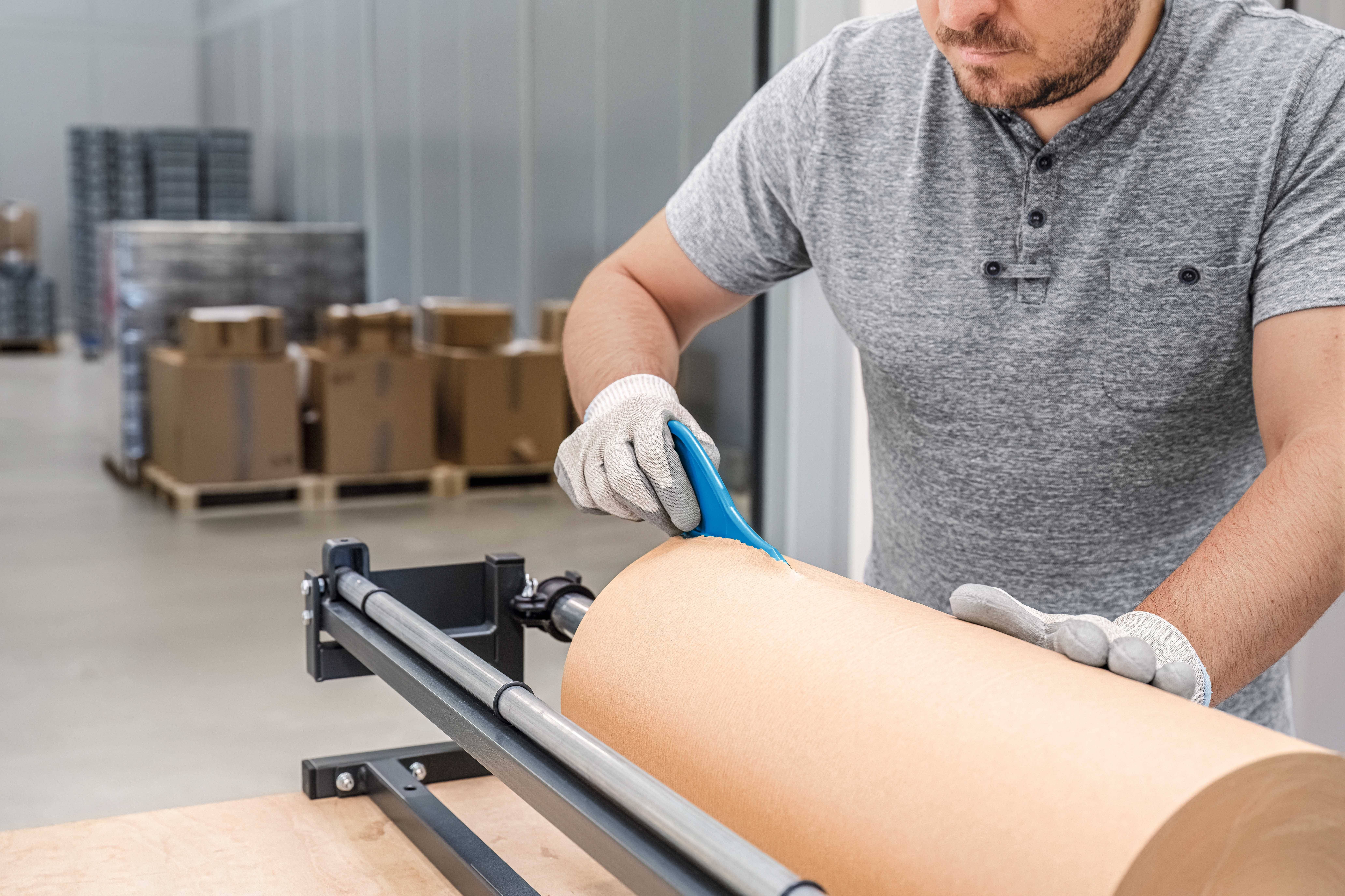 A man is cutting a roll of packaging paper on a machine in a warehouse. Cardboard boxes are stacked in the background.