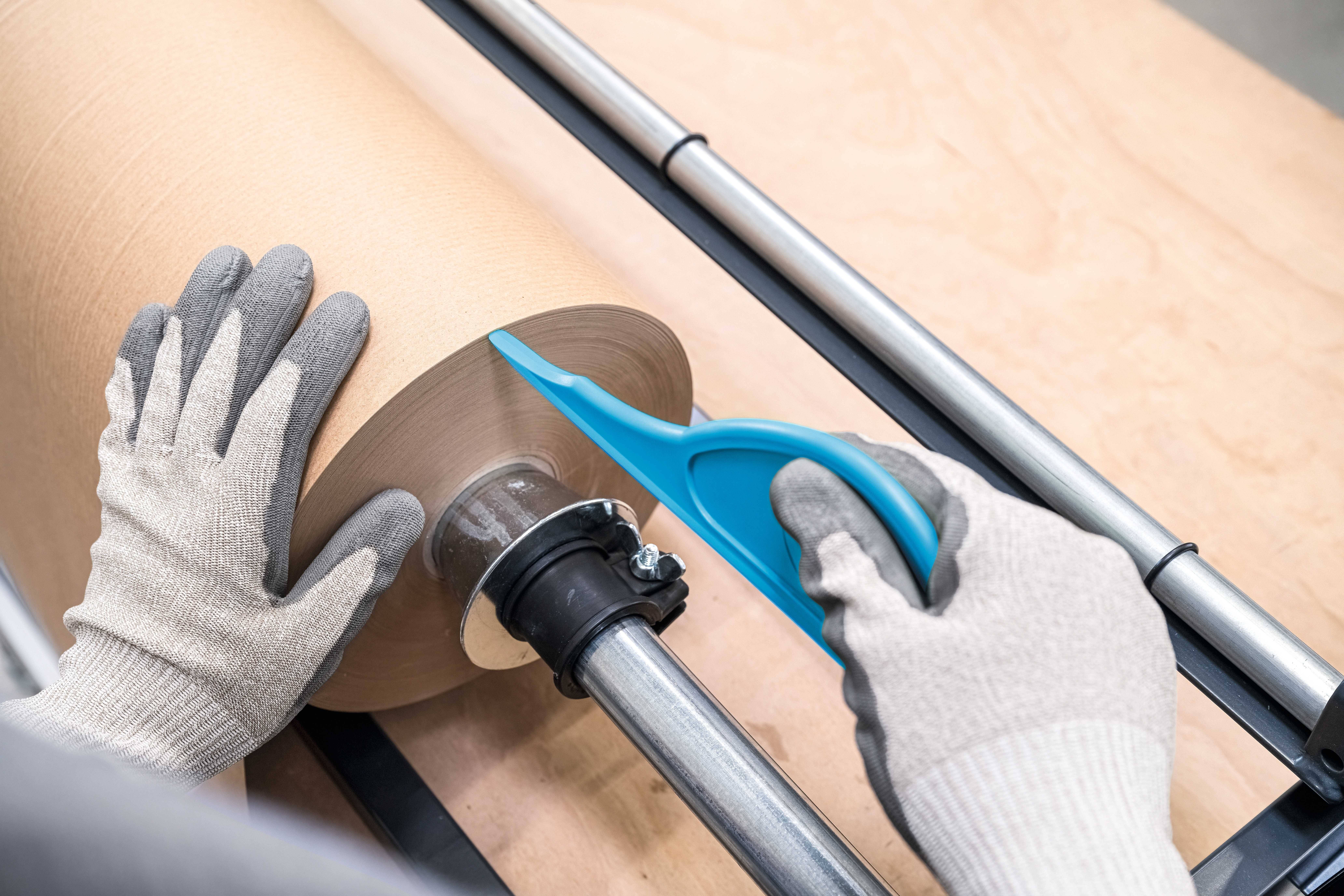 A person is cutting a large paper roll core with a blue knife. They are wearing grey gloves for protection.