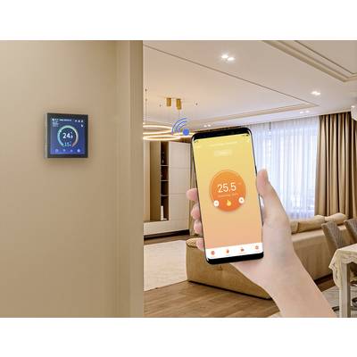 A hand is holding a smartphone displaying the control of a room thermostat. The room is furnished in a modern style with large windows.