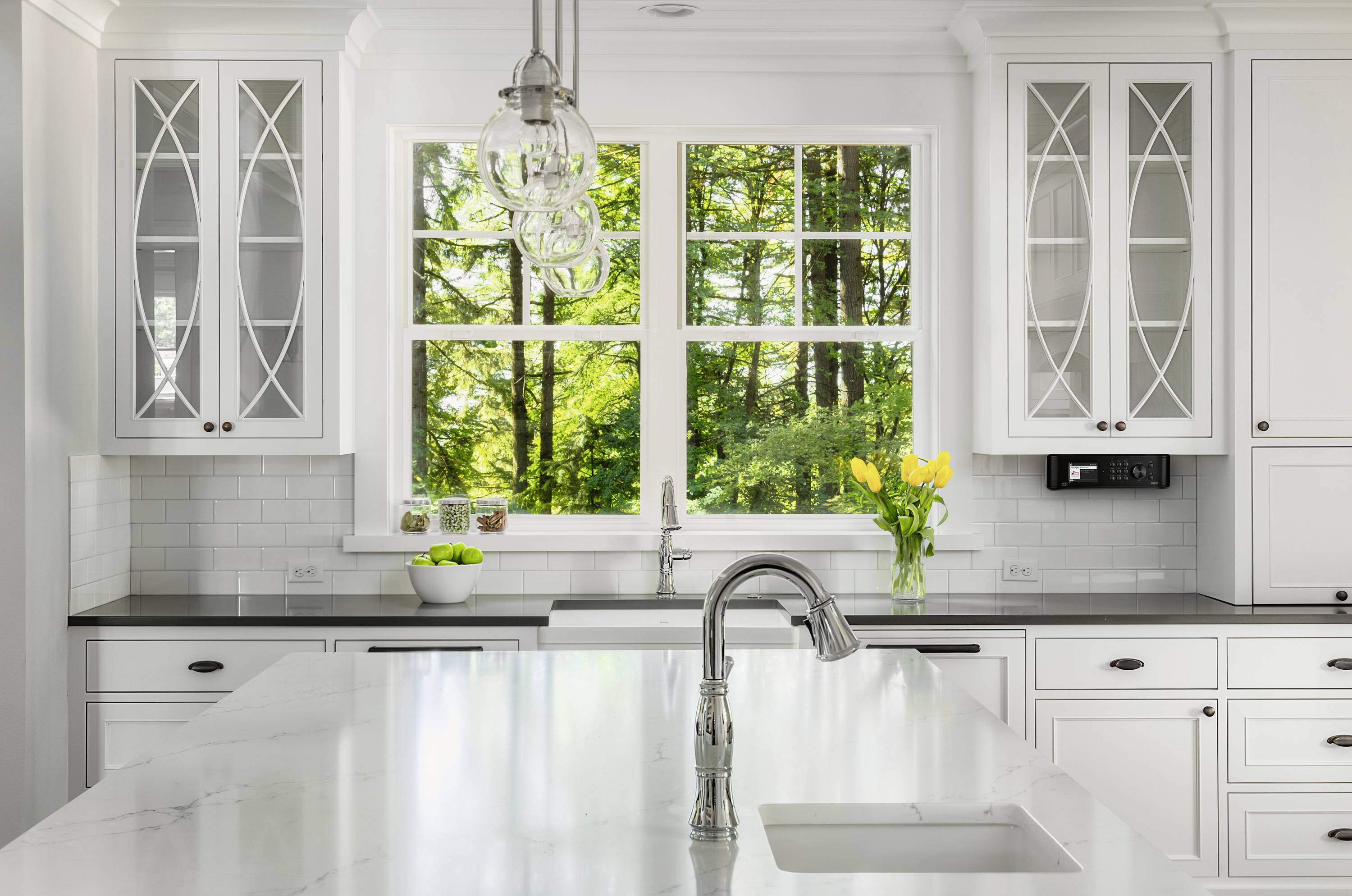 Bright, modern kitchen with white cabinets, marble island and large window offering a view of green trees. Yellow tulips on the worktop.