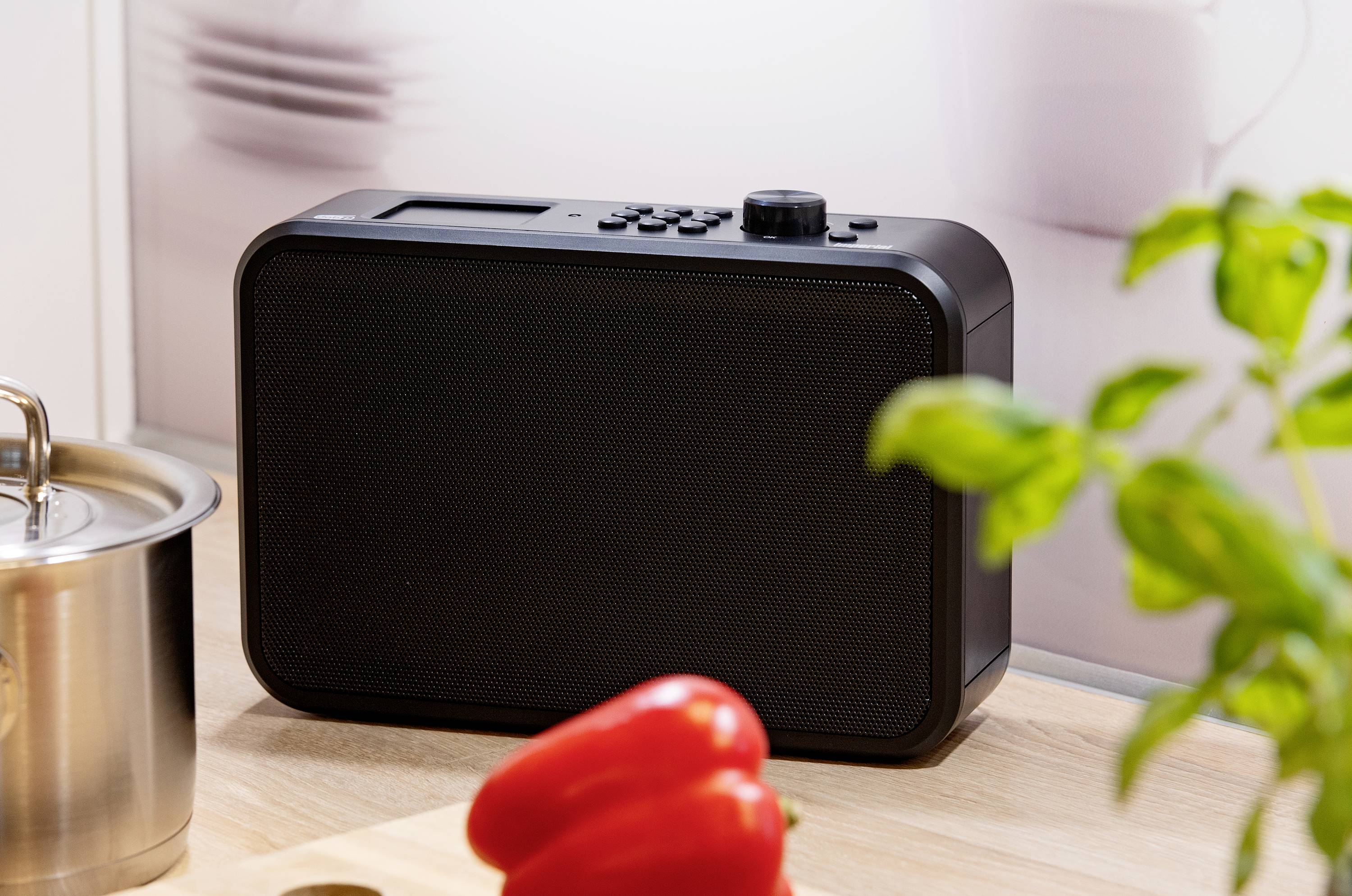 A black radio sits on a kitchen worktop next to a pot, red peppers, and basil.