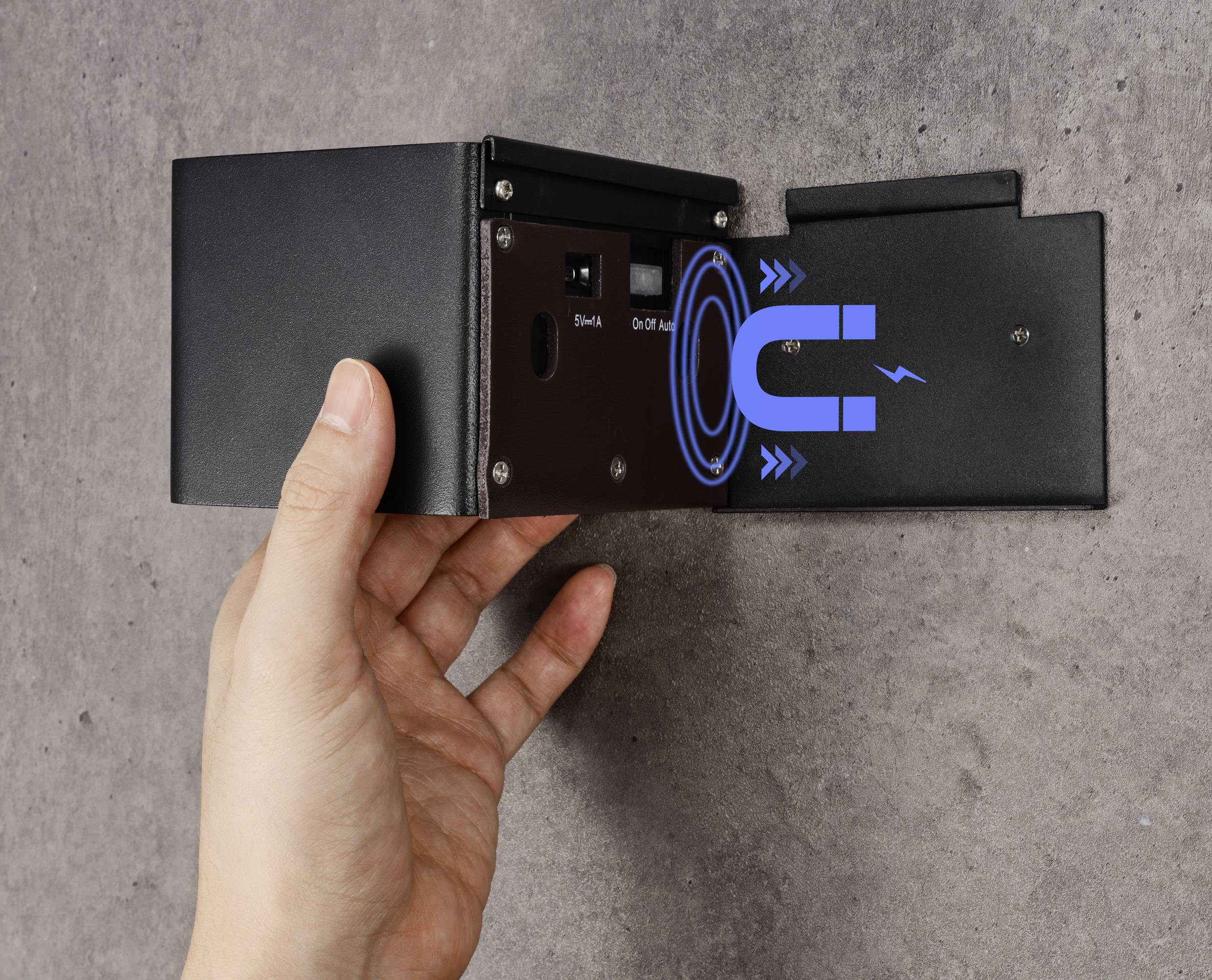 A hand is holding a black technical device with a magnet symbol on the side. The device is mounted on a grey concrete wall.