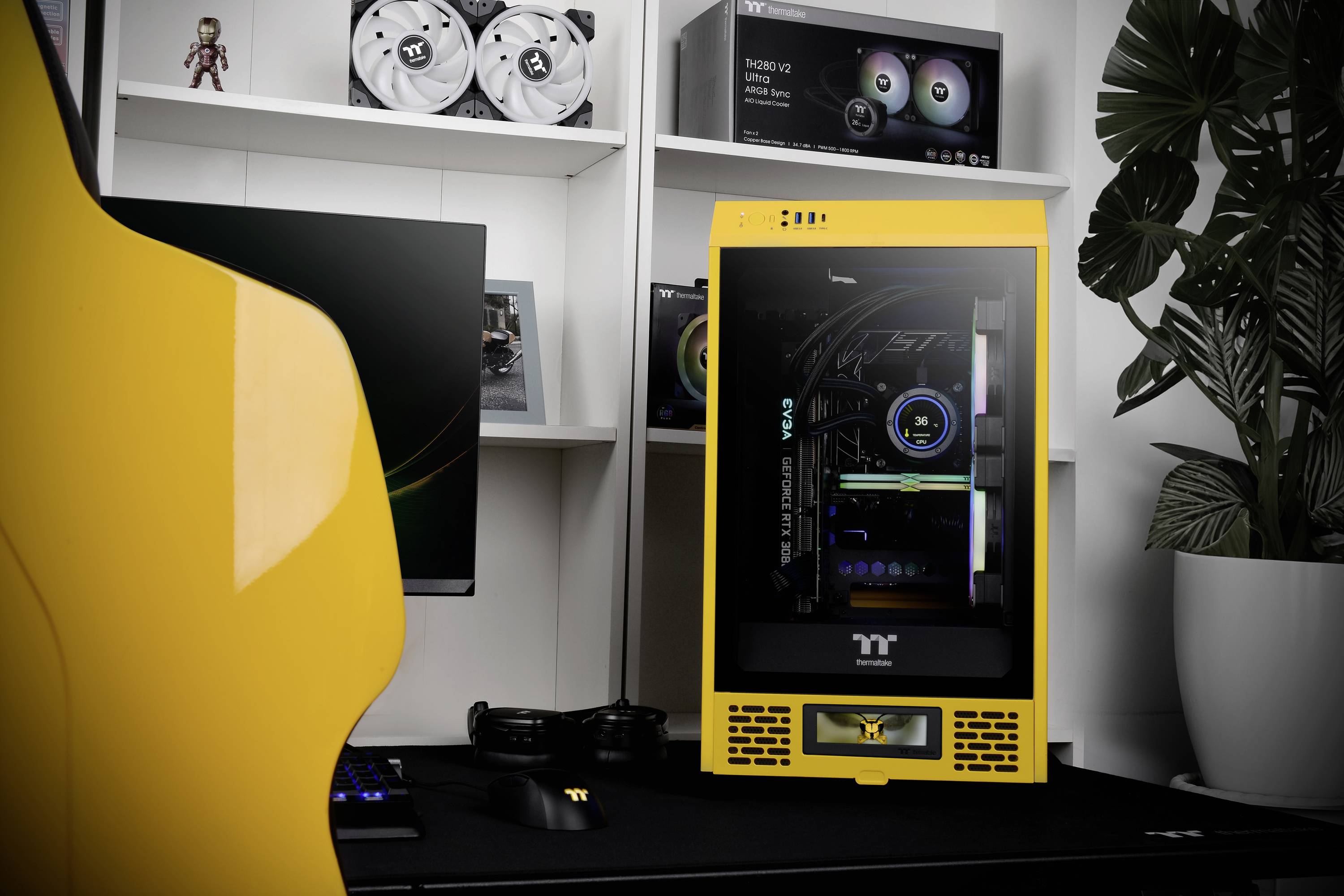 A yellow gaming PC with a transparent case reveals its internal hardware. In the background, a white shelf with computer components and a green plant can be seen.
