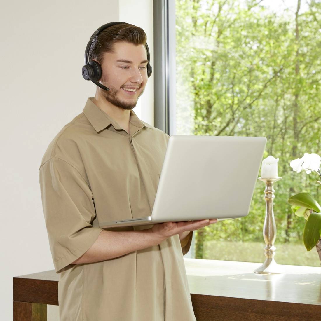 Man wearing a headset working on a laptop, smiling at the screen. In the background, a window with a view of trees.