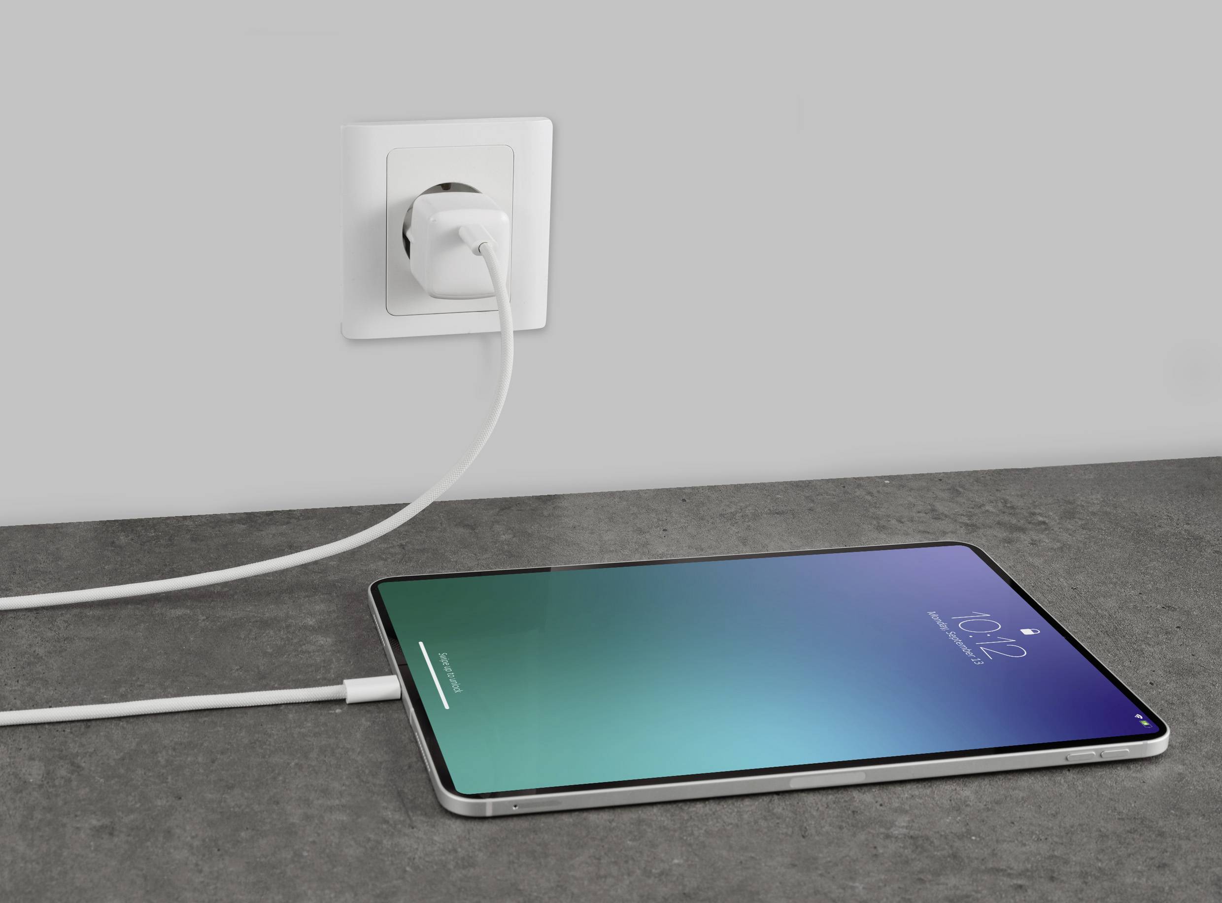 A tablet is lying flat on a table and is being charged via a cable connected to a wall socket.