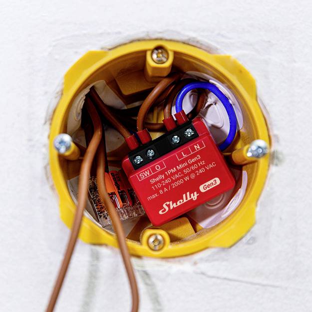 'Close-up of a Shelly 1PM Mini relay, installed in a yellow wall socket, with visible wiring. Label shows specifications.'