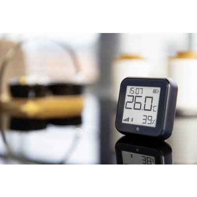 A digital thermo-hygrometer displays 26.0°C and 39% humidity, time 15:07. Household items blurred in the background.