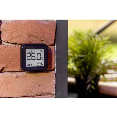 A digital thermometer on a brick wall displays 26°C and 39% humidity. A plant is blurrily visible in the background.