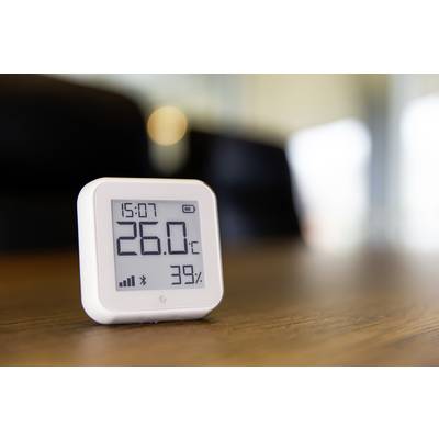 A digital thermometer displays 26.0 °C and 39% humidity on a wooden table, with a room in the background.