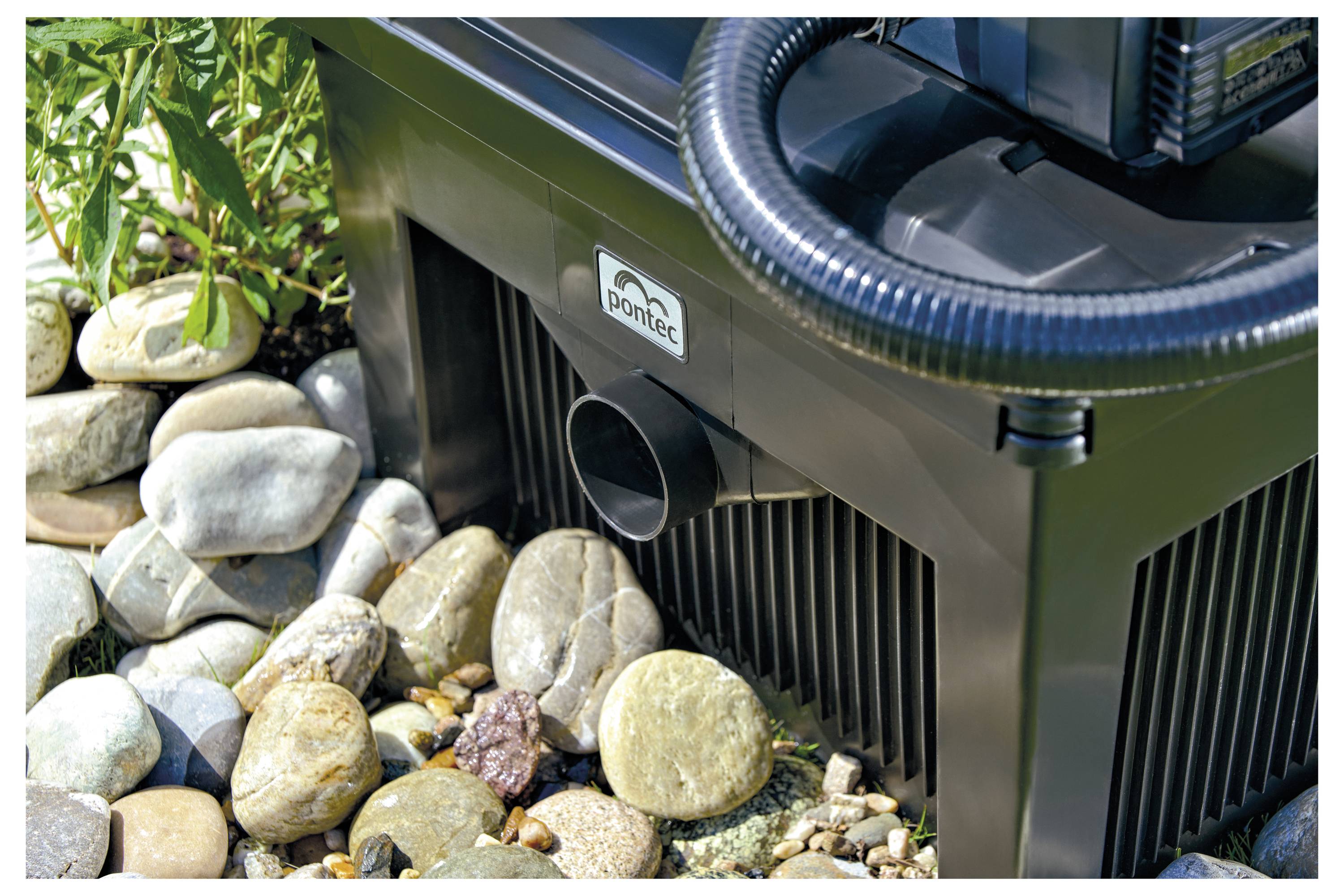 A water pump is placed on smooth, rounded stones. A black hose is connected to the pump, with green foliage visible on the left.