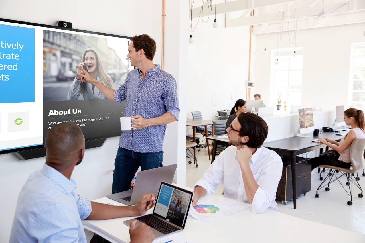 A man is presenting to colleagues in front of a screen in a modern office. Employees are working on computers in the background.