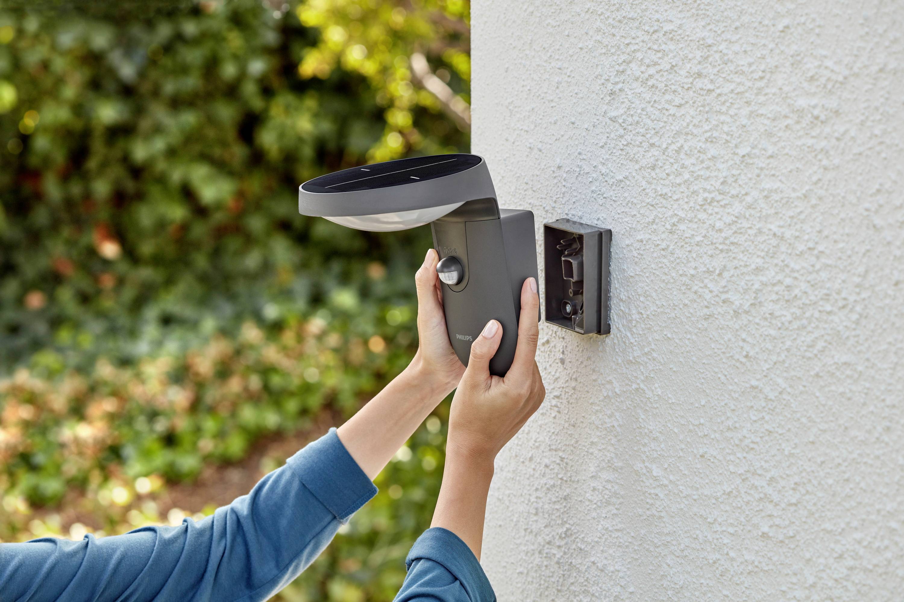 A person is installing an exterior light on a wall. Blurred green foliage can be seen in the background.