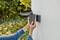 A person is installing an exterior light on a wall. Blurred green foliage can be seen in the background.