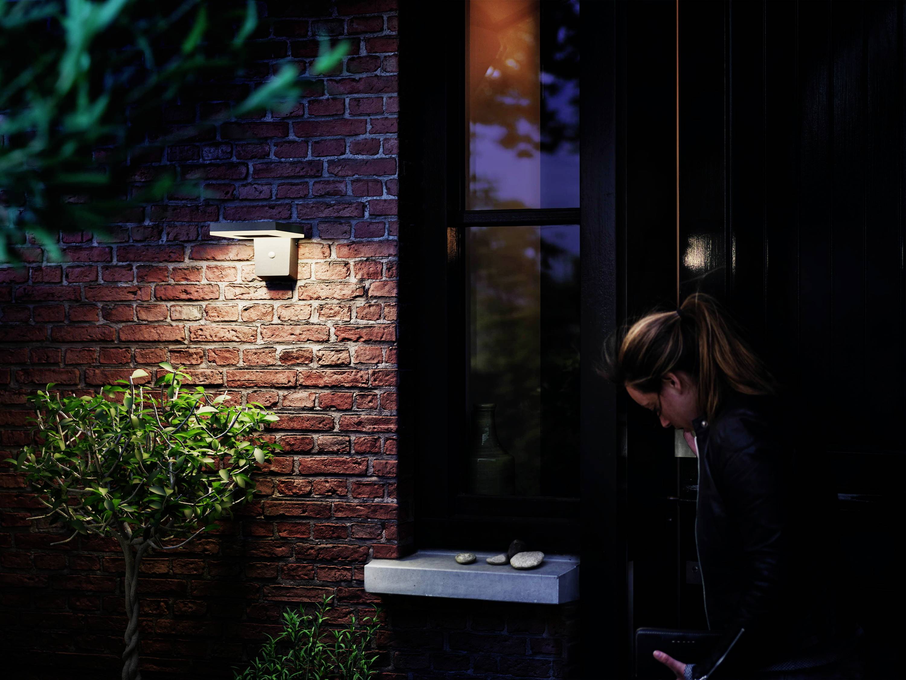 A woman opens a door in a brick house at nightfall. An exterior light illuminates a small tree and the entrance area.