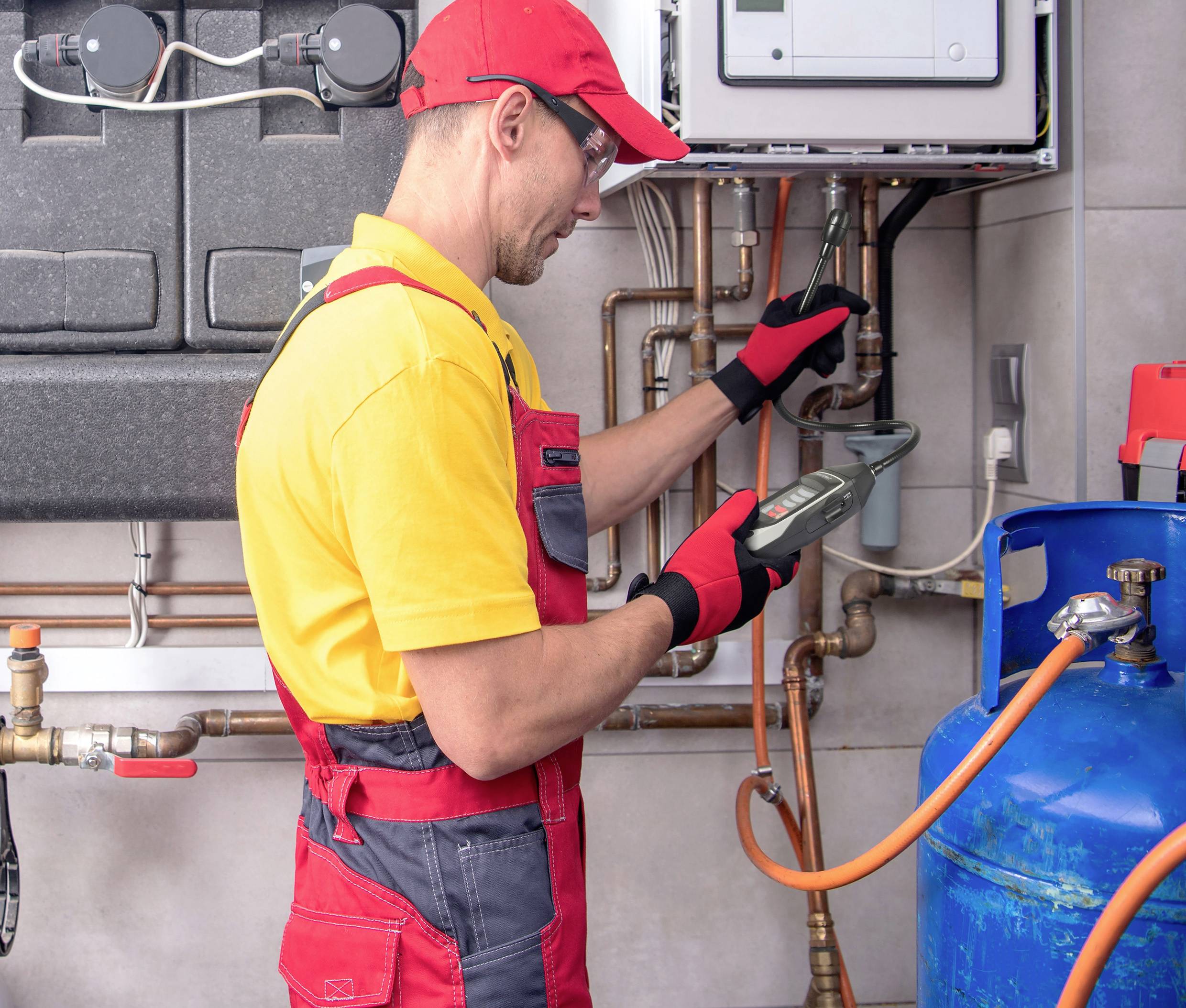 A tradesman in yellow and red workwear is checking a gas pipeline with a measuring device, surrounded by pipes and a blue gas tank.