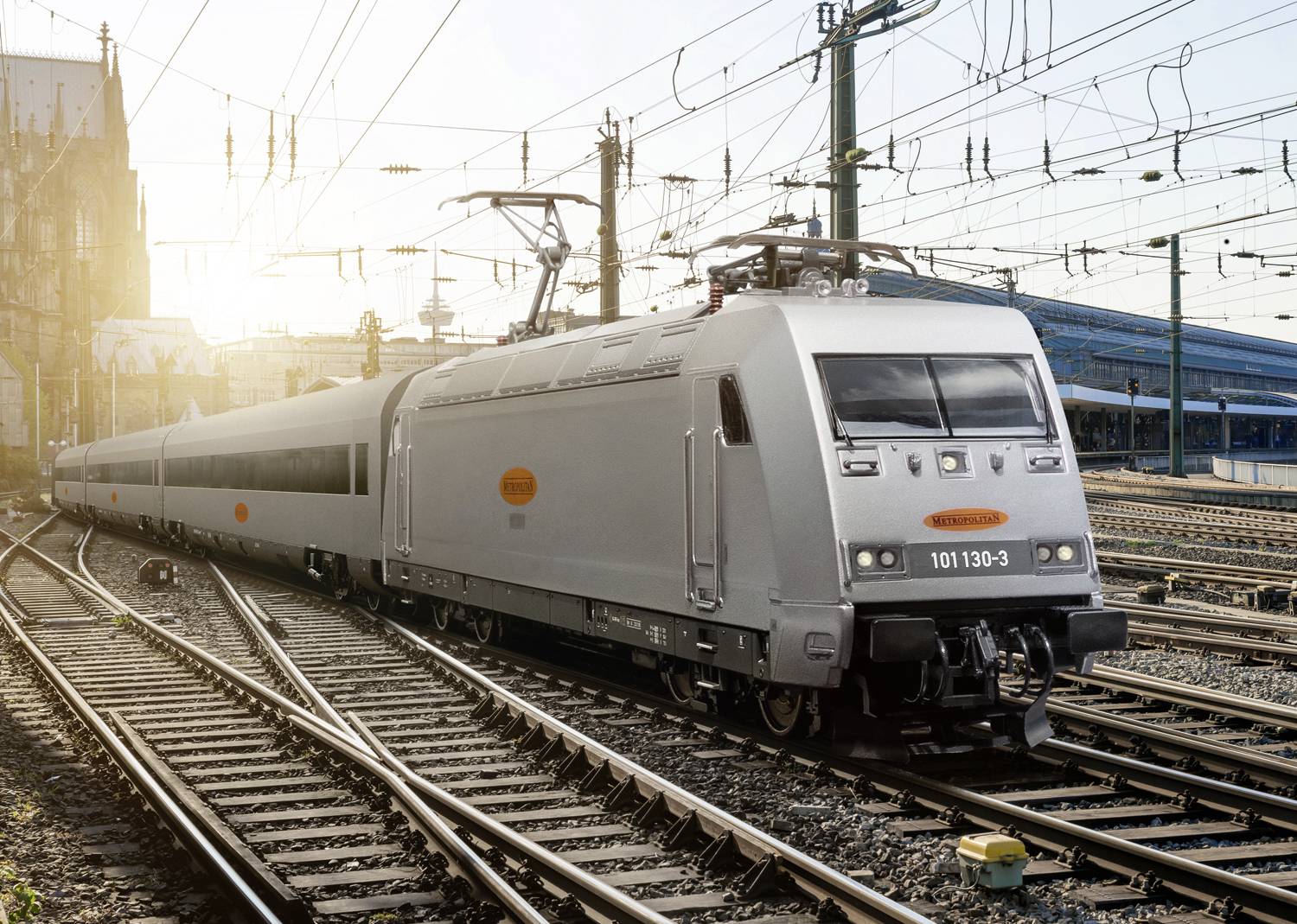 A modern high-speed train travels past a station at sunset, with overhead power lines visible in the surrounding area.