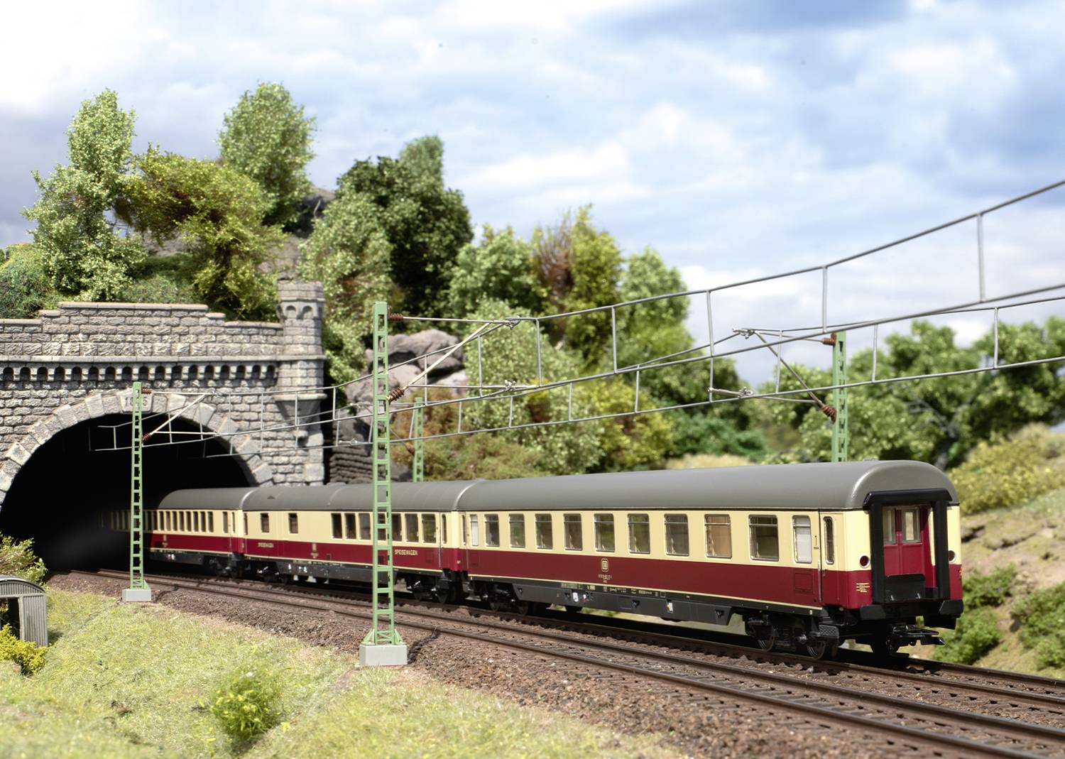 A train emerges from a tunnel in a rural setting, surrounded by green vegetation and clouds in the sky.