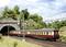 A train emerges from a tunnel in a rural setting, surrounded by green vegetation and clouds in the sky.