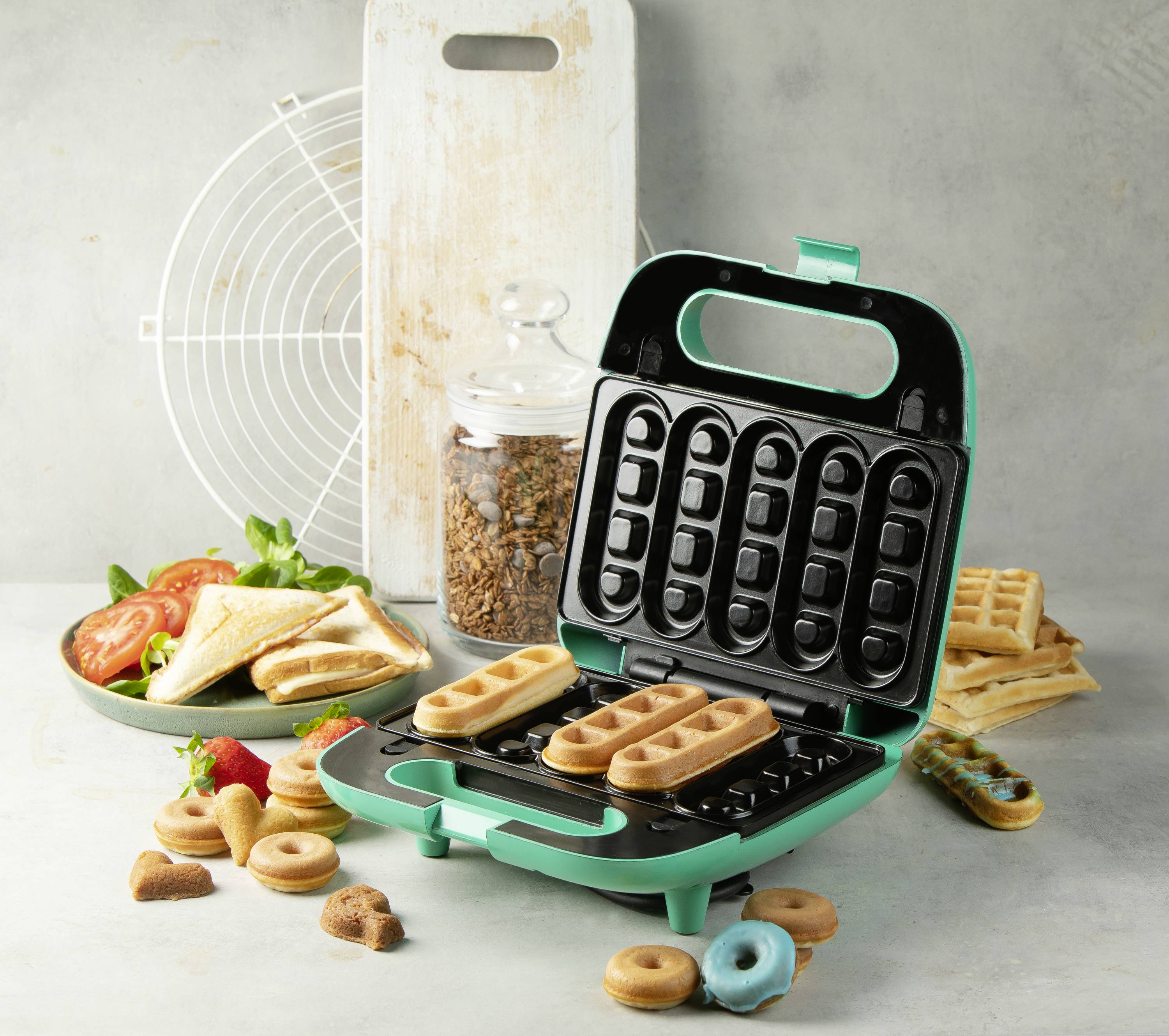 A green waffle machine with fresh waffles and accompanying pastries, alongside a plate of sandwiches and tomatoes.