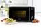 A black microwave sits on a white table in a kitchen. Beside it is a plate of green pasta and cherry tomatoes.